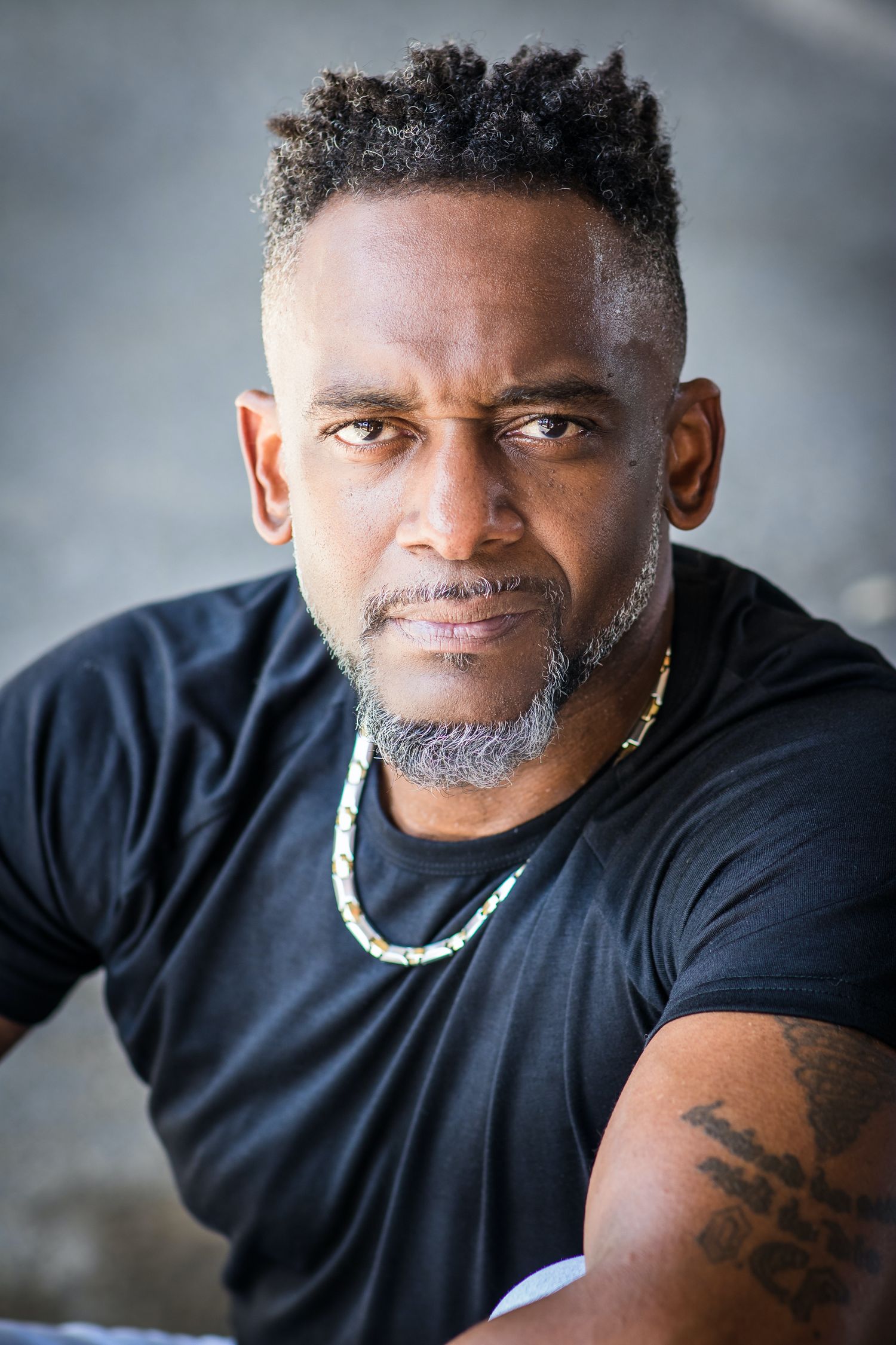 Professional studio portrait of a man in black t-shirt wearing a gold chain necklace.
