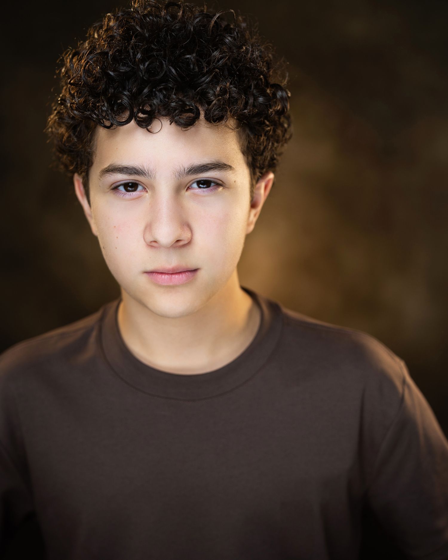 Young teen boy with curly dark hair wearing a brown shirt poses for a professional headshot photo.