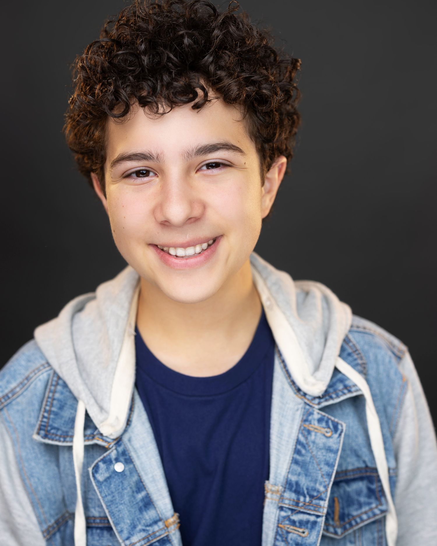 Smiling teenage boy with curly hair wearing a denim jacket over a hoodie and navy shirt, professional headshot.