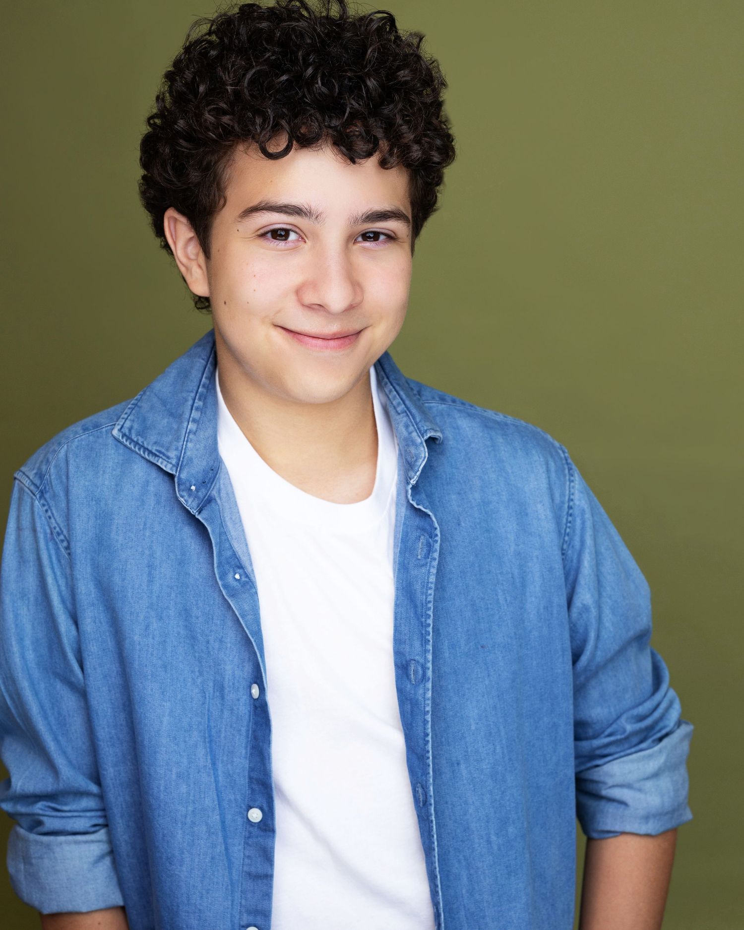 Smiling teenage boy with curly dark hair wearing a denim shirt over a white t-shirt against a green background.