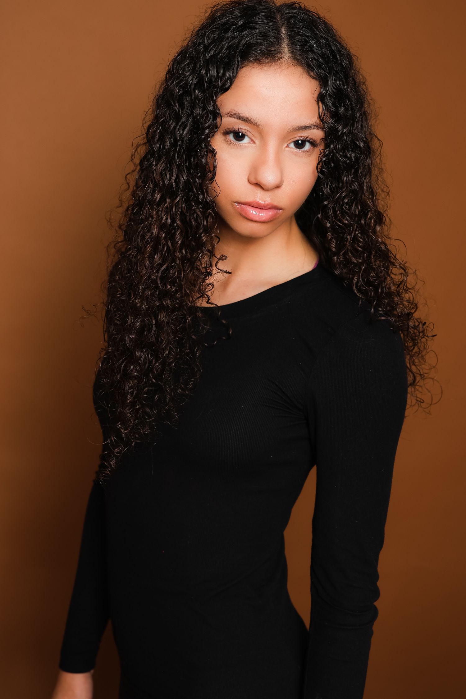 Portrait of a person with long curly dark hair wearing a black long sleeve top against a brown background.