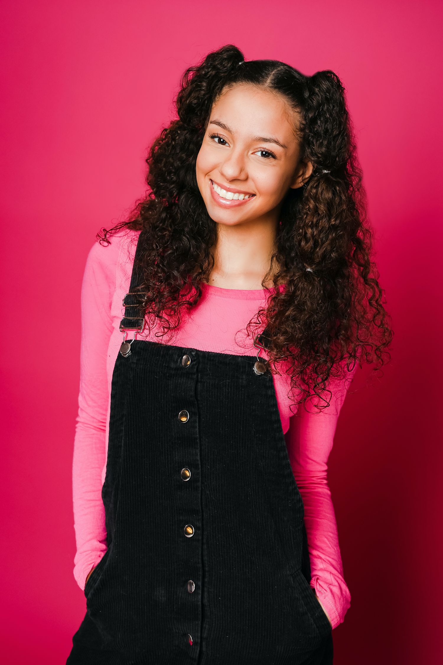 Young woman with curly hair wearing pink long sleeve top and black overalls poses against bright pink background.