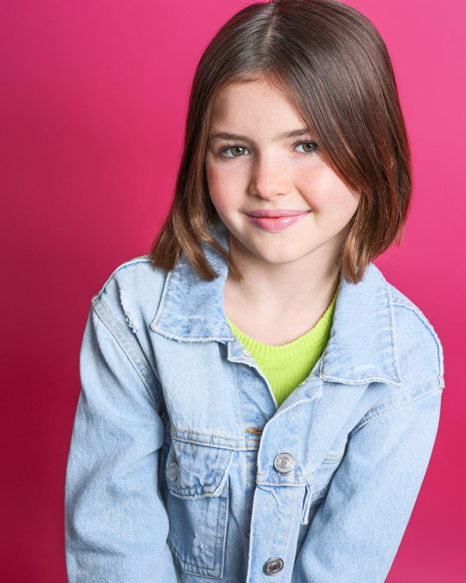 Person with straight brown hair poses against pink background wearing denim jacket and neon yellow top.