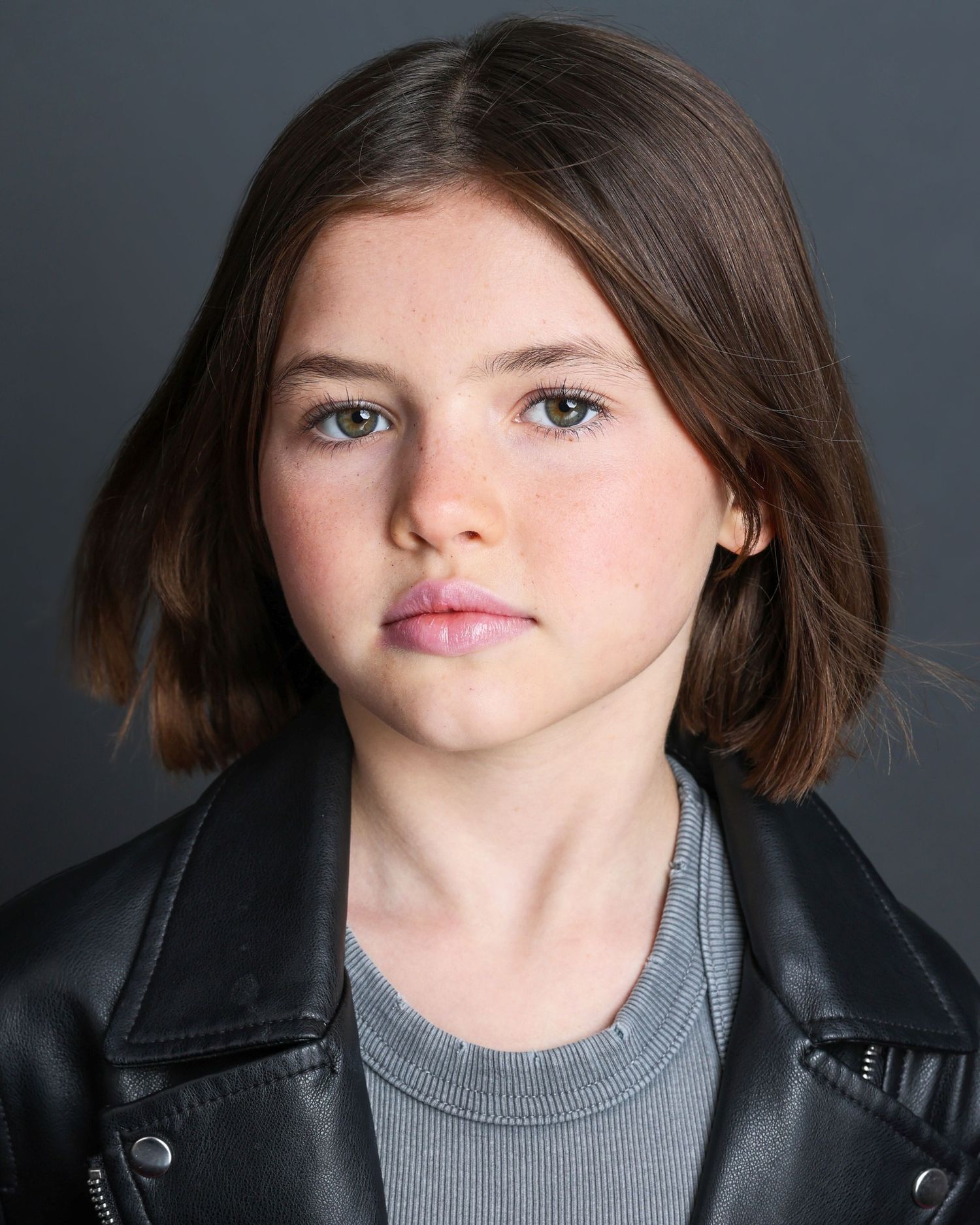 Professional headshot portrait of someone in a black leather jacket and gray shirt against a dark background.