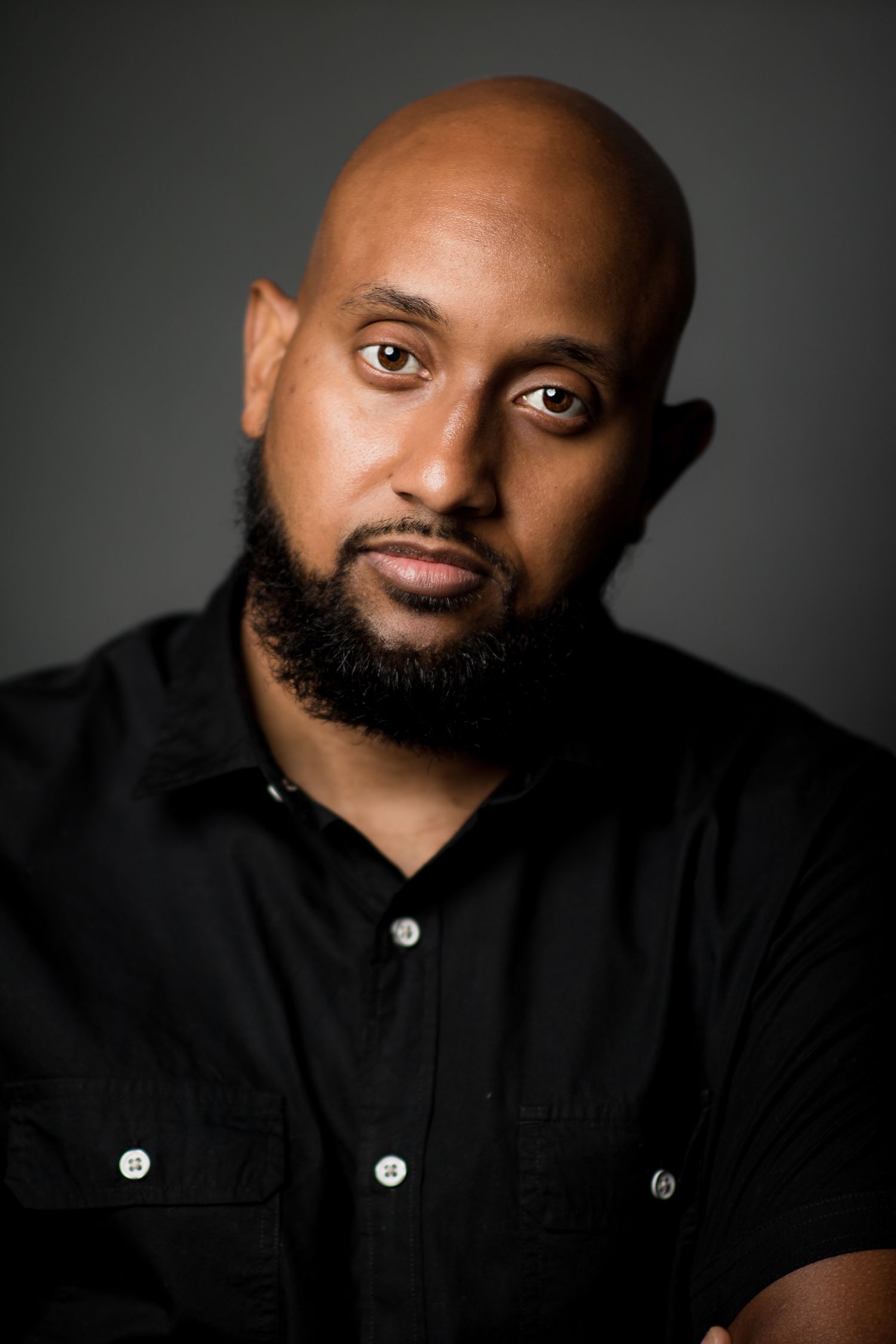 Professional headshot of a person with beard wearing black button-up shirt against dark background.
