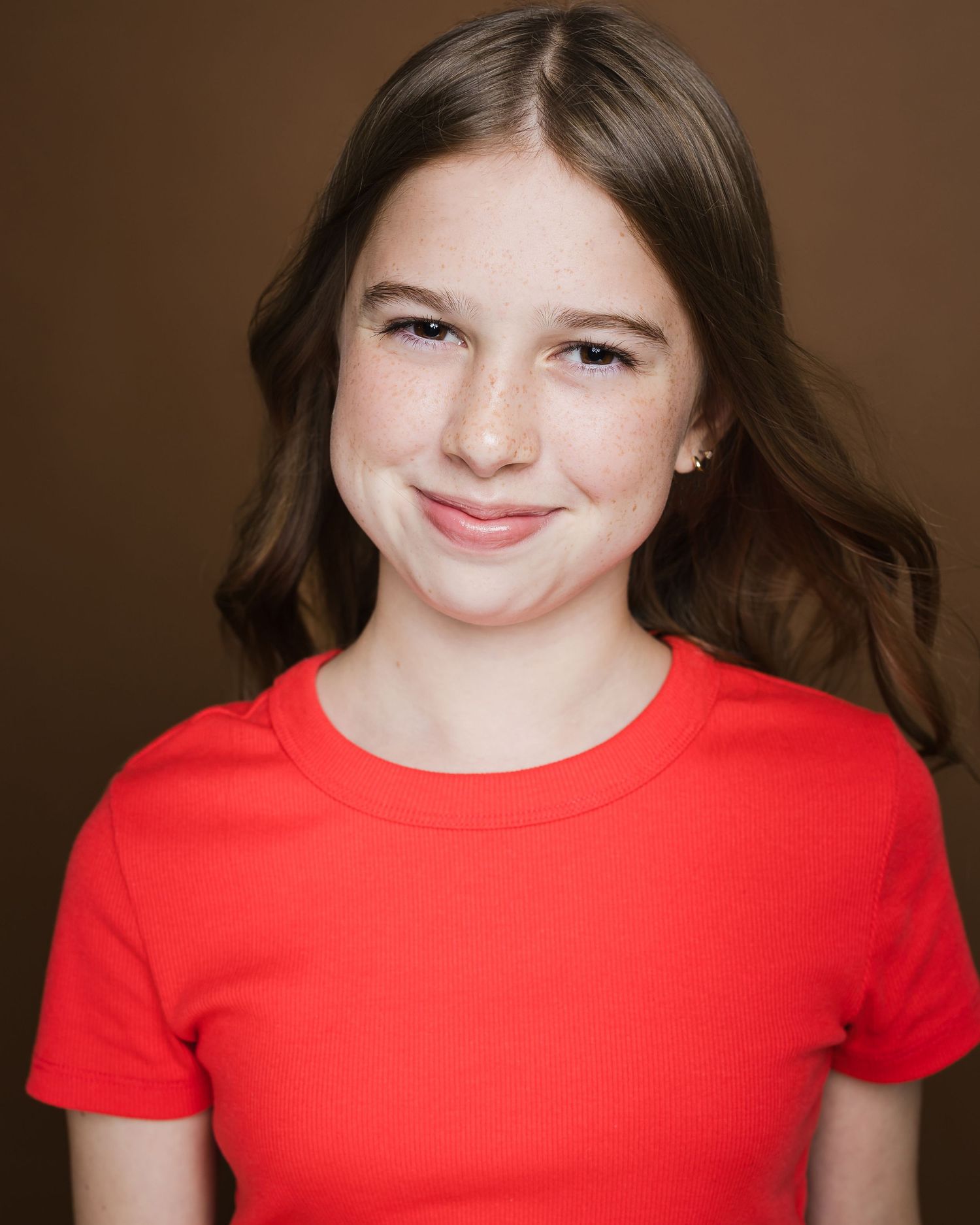 Person in red t-shirt smiles kindly against dark brown background in headshot portrait series.
