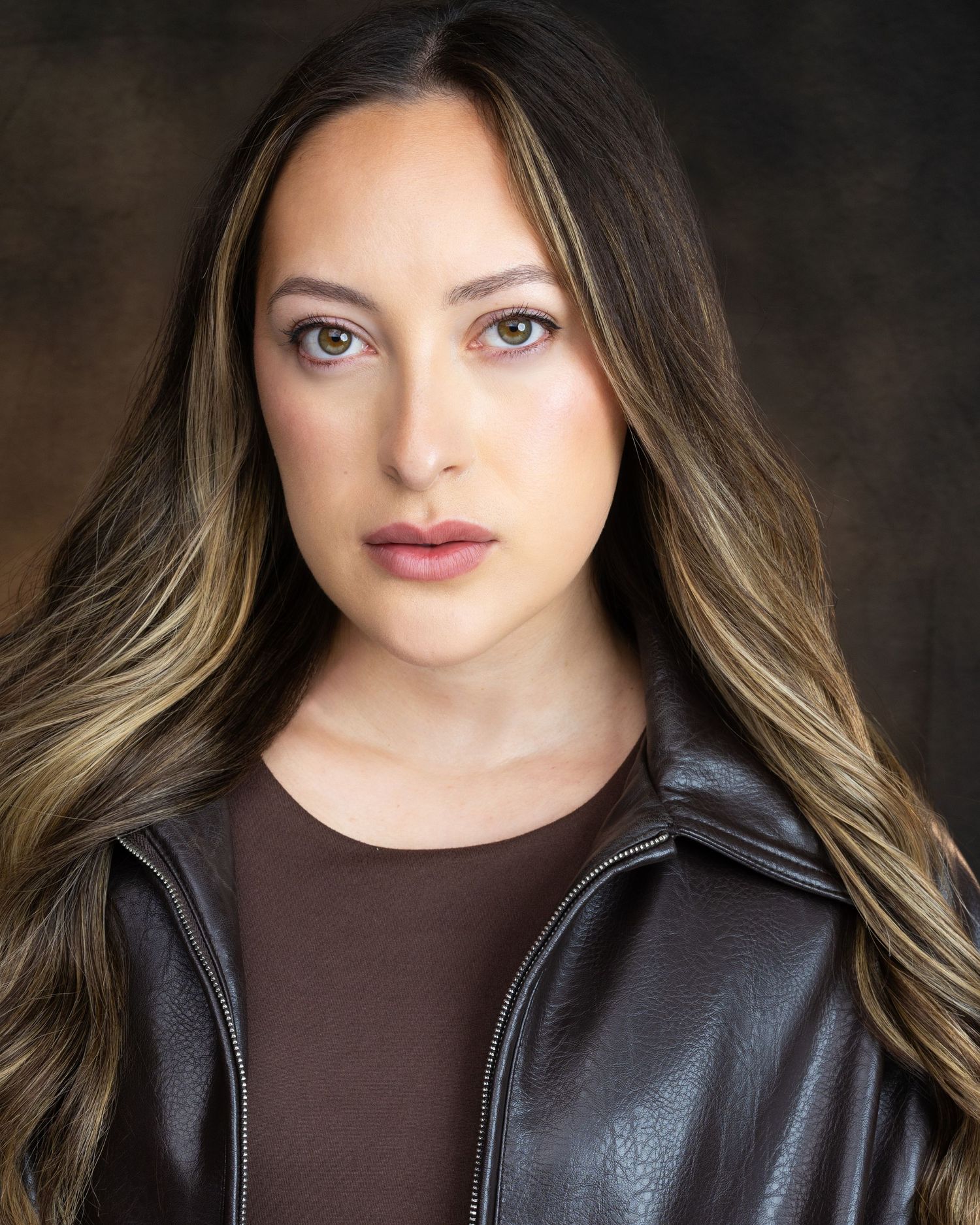 Young woman with long highlighted hair wearing a brown leather jacket poses for a professional headshot.