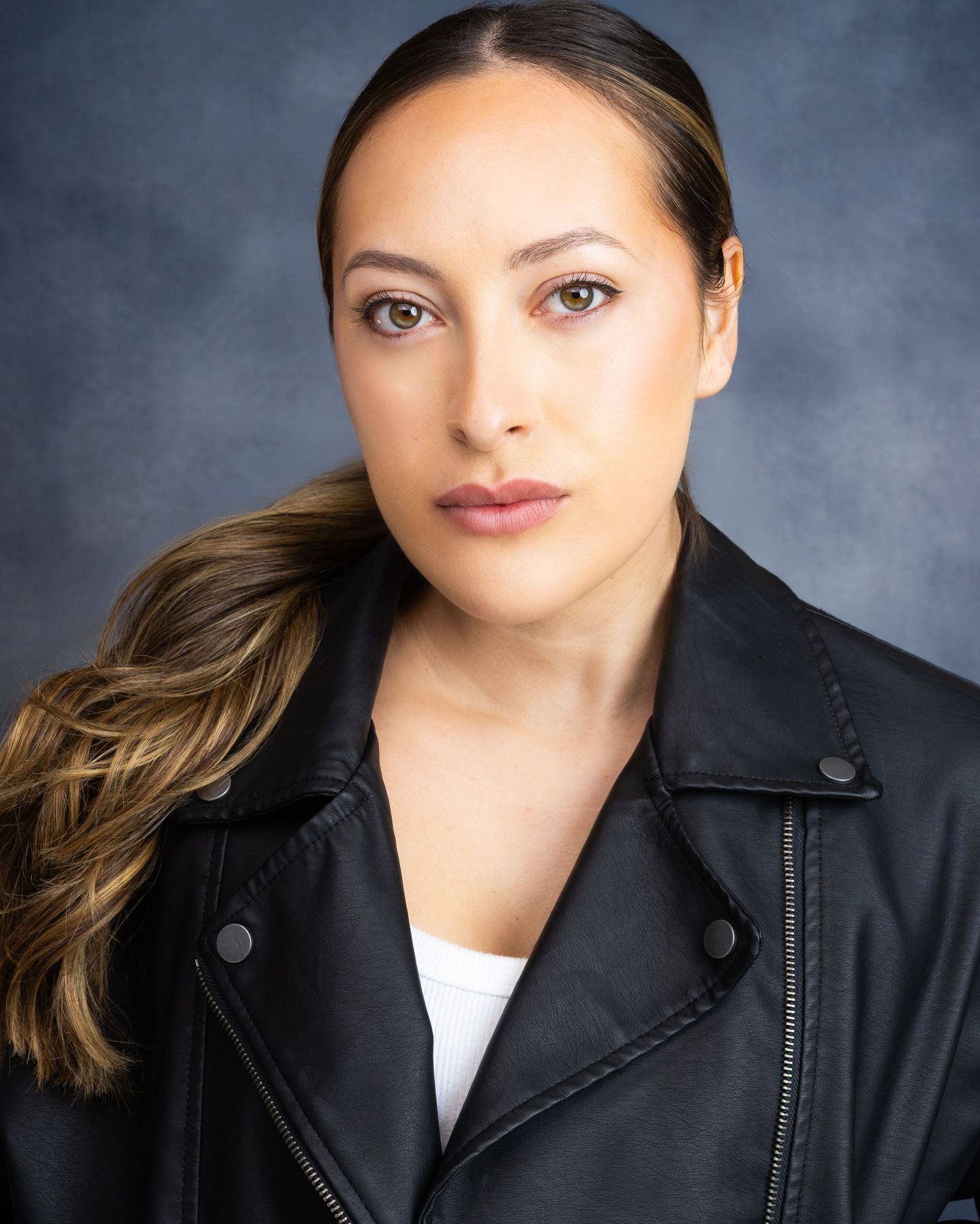 Professional headshot of a young woman in a black leather jacket with highlighted hair against a gray backdrop.