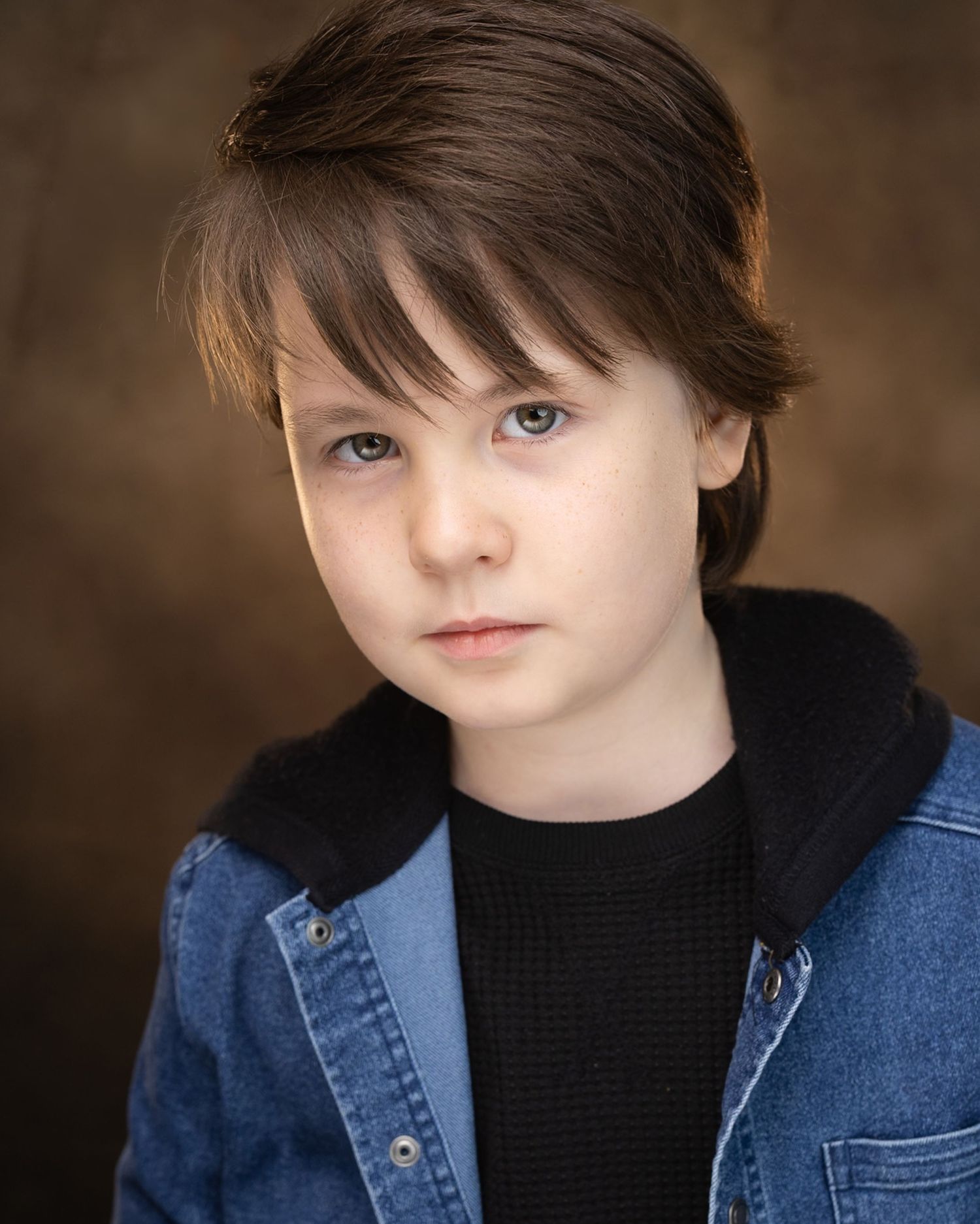 Young boy with brown hair wearing a denim jacket over a black sweater, posing for a professional headshot photo.