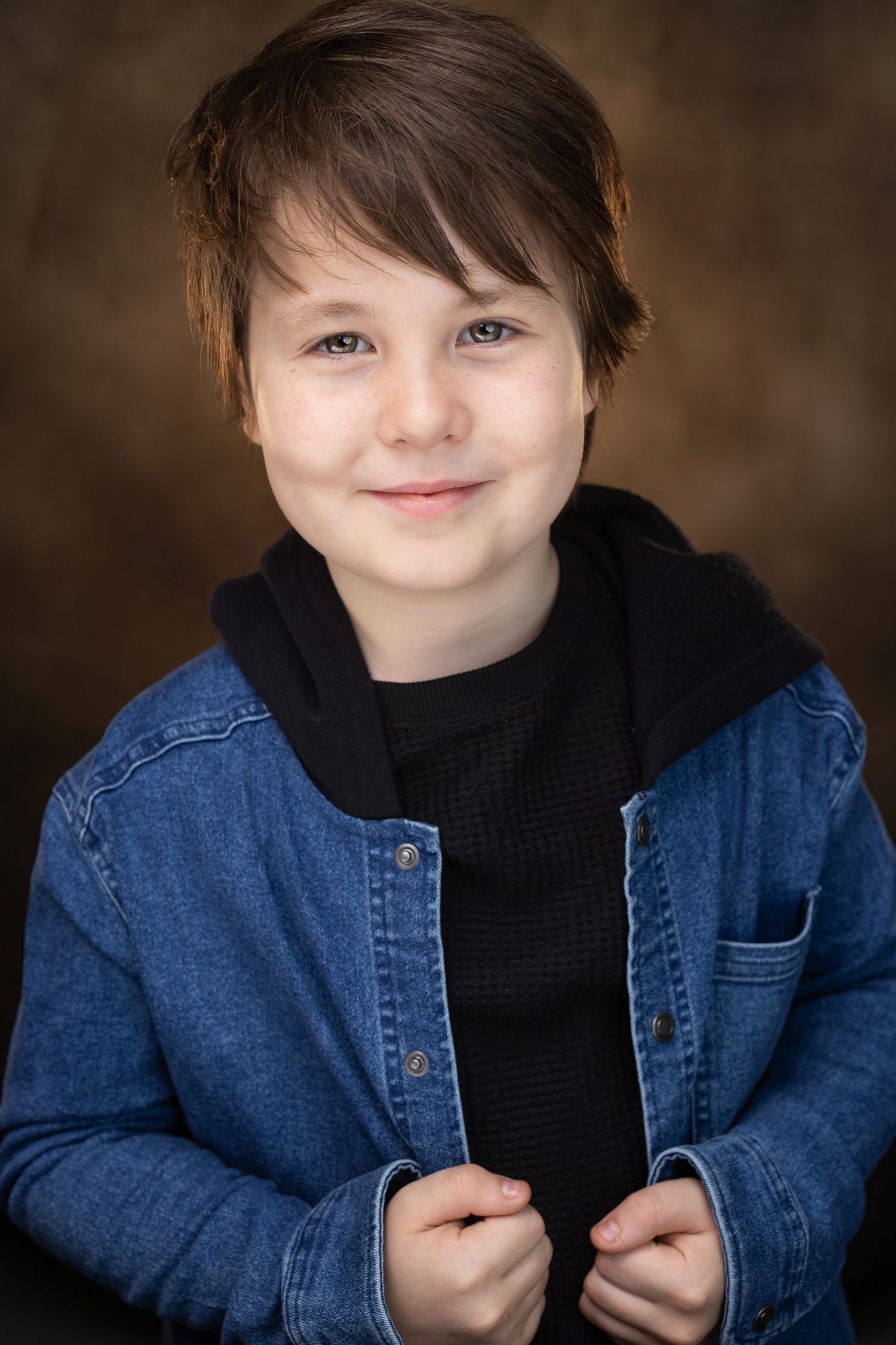 Smiling young boy in a blue denim jacket over a black hoodie, posing against a brown studio background.