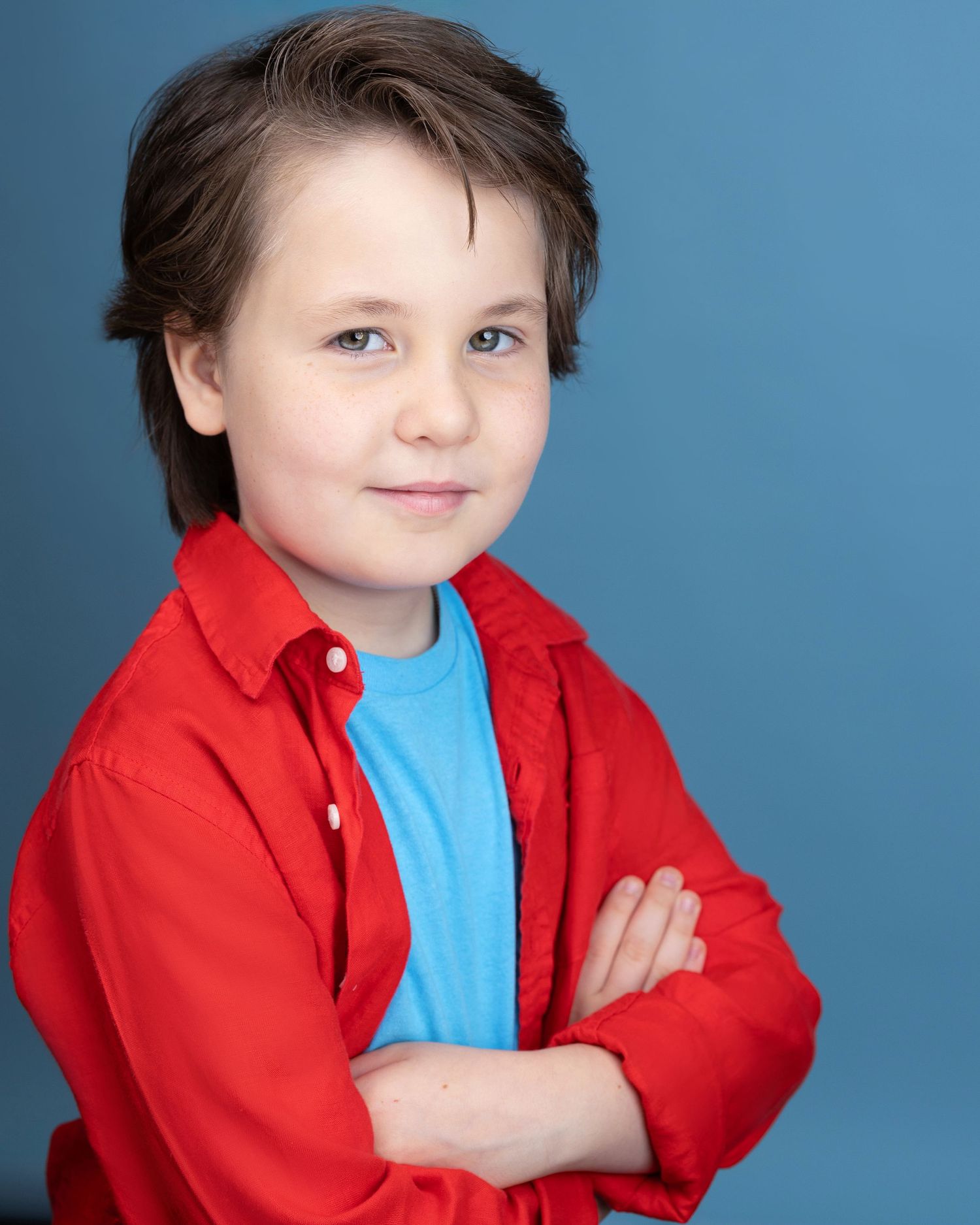 Young boy with brown hair in red shirt and blue tee, arms crossed, smiling against blue background.