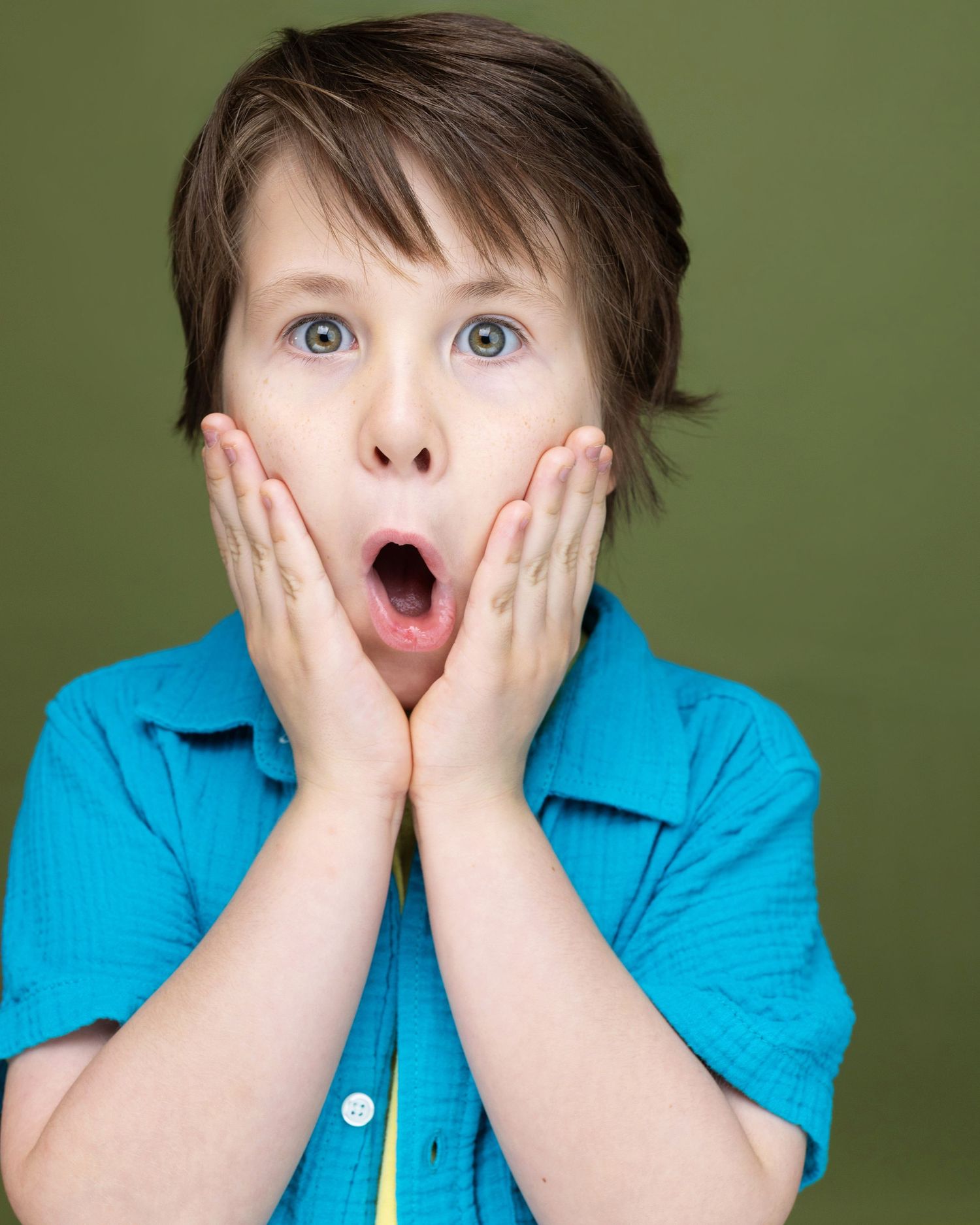 A surprised young boy with blue eyes holds his cheeks with both hands, mouth open wide, wearing a turquoise shirt.