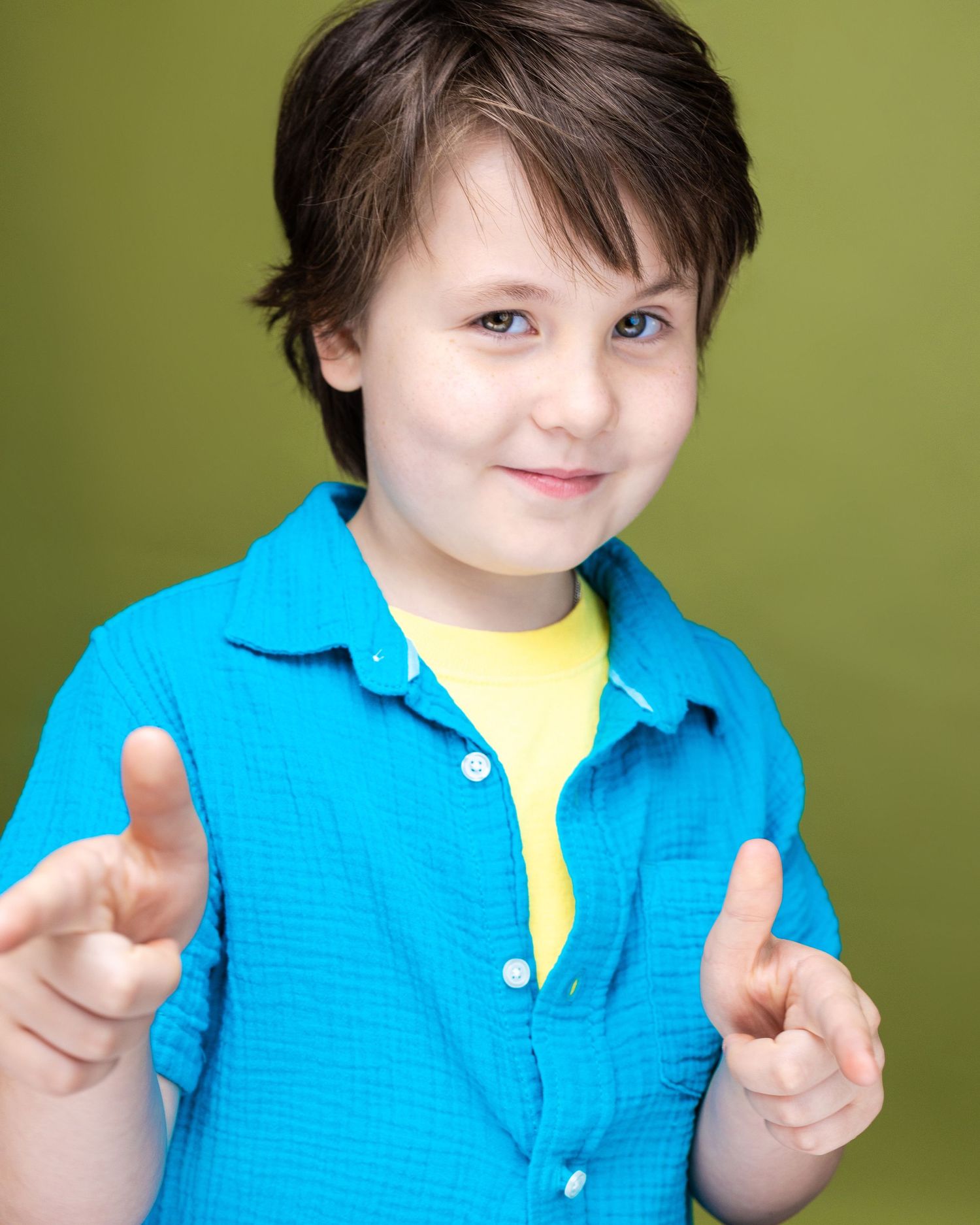 A cheerful young boy in a blue shirt and yellow tee points finger guns at the camera against a green background.