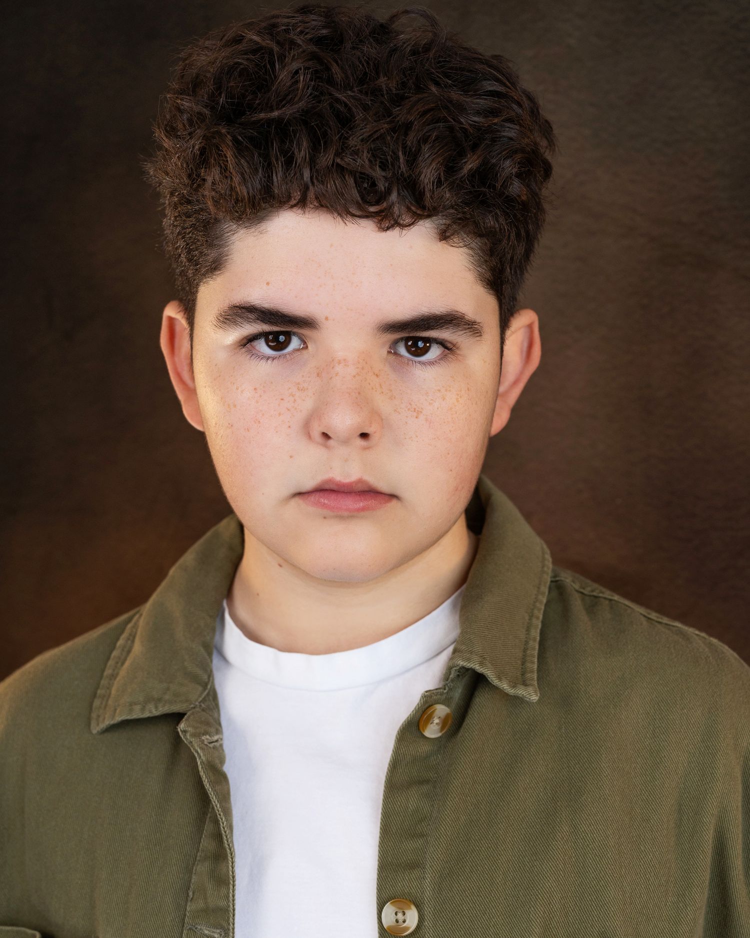 Young teen with curly dark hair and freckles wearing a green jacket over white shirt in a professional headshot.