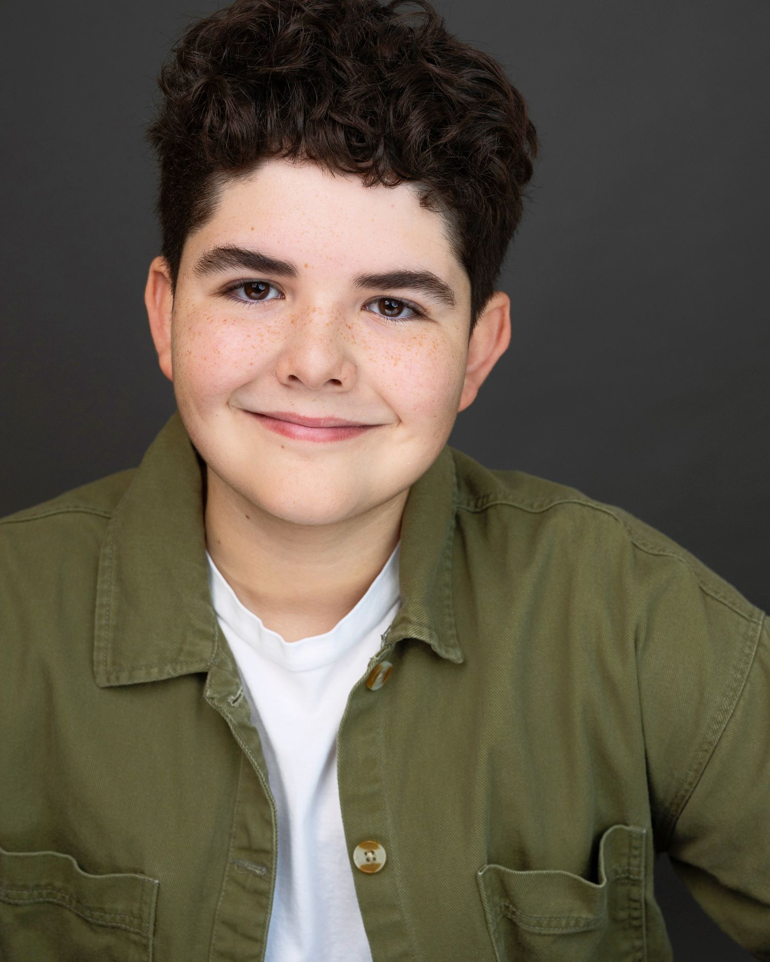 Young boy with curly dark hair wearing a green jacket and white shirt, smiling in a professional headshot.