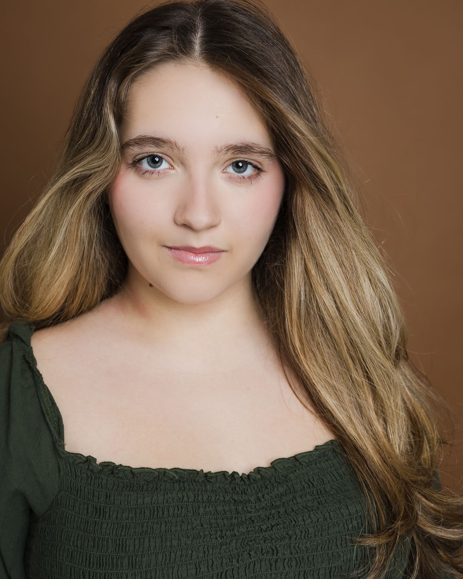 Portrait against brown backdrop showing long blonde ombre hair and forest green smocked top.