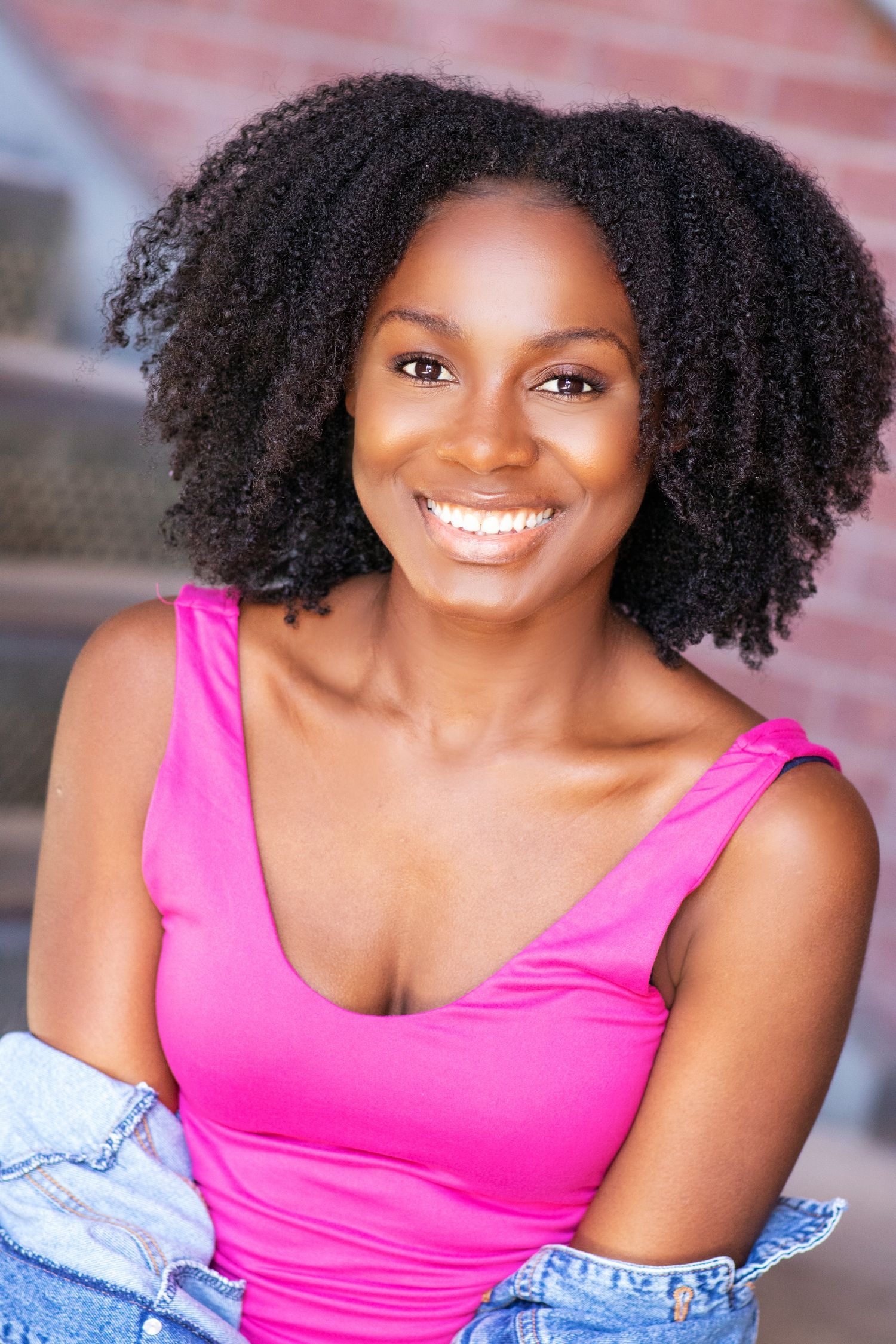 Joyful headshot of a woman with natural curly hair wearing a bright pink tank top and denim jacket.