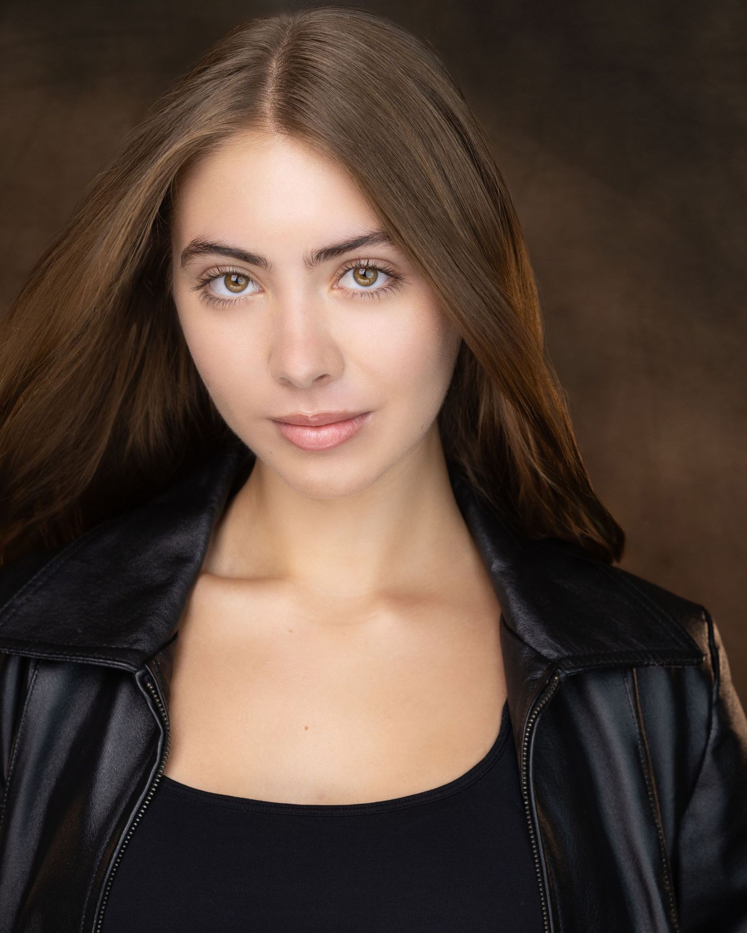 Young woman with long brown hair and green eyes wearing a black leather jacket, professional headshot photo.