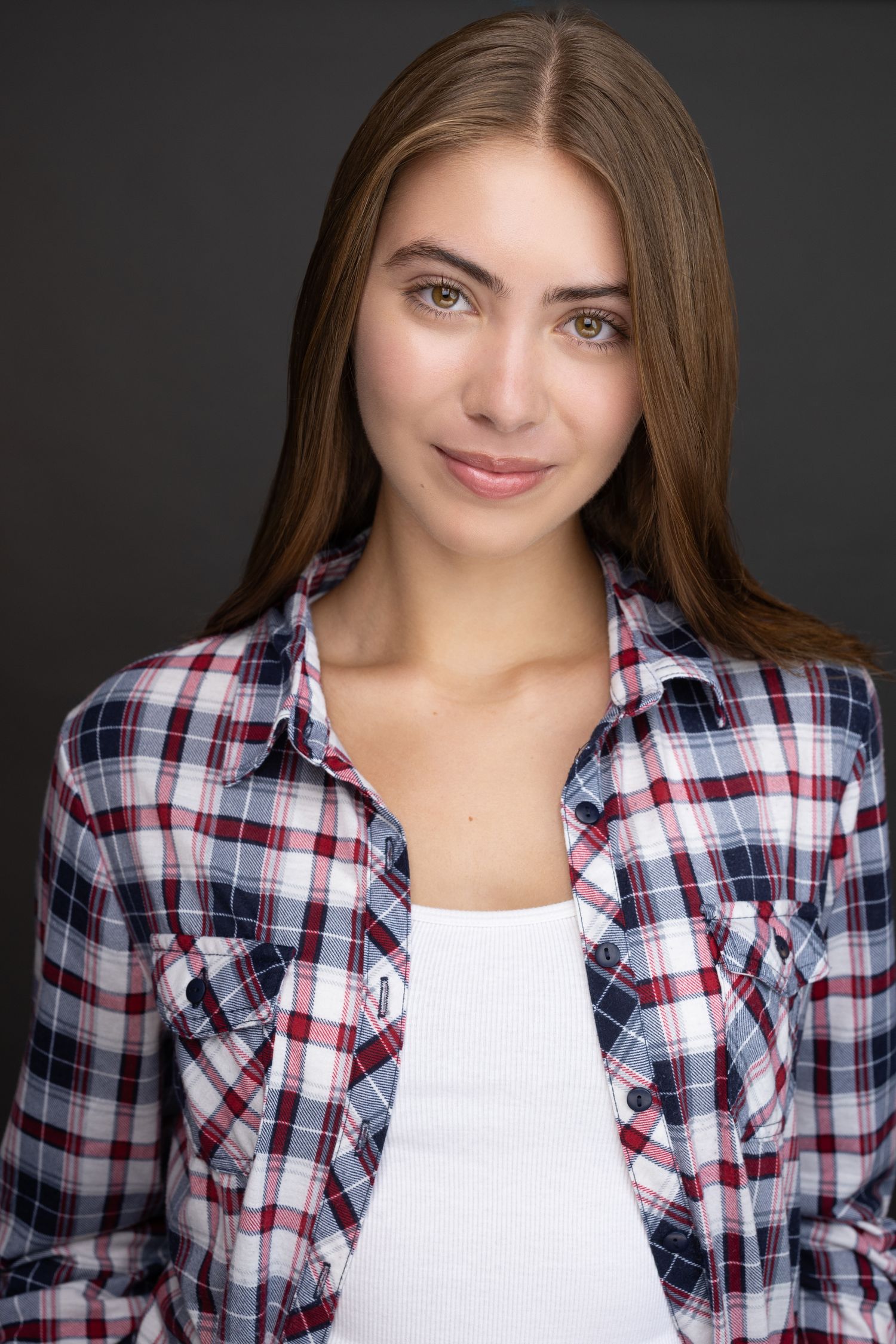 Young woman with long brown hair wearing a plaid flannel shirt over white top, smiling against dark background.