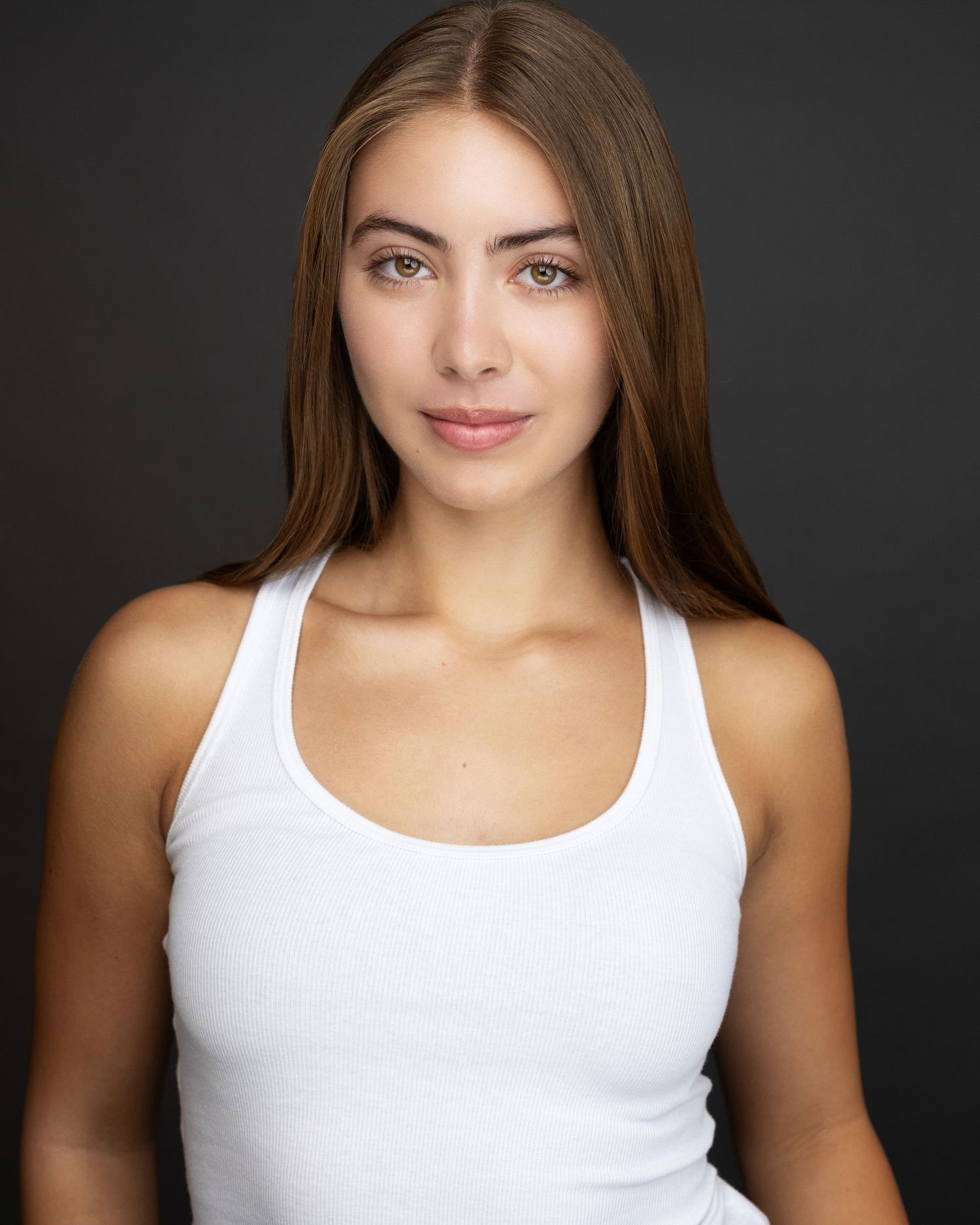Young woman with long brown hair wearing a white racerback tank top poses against a dark gray background.