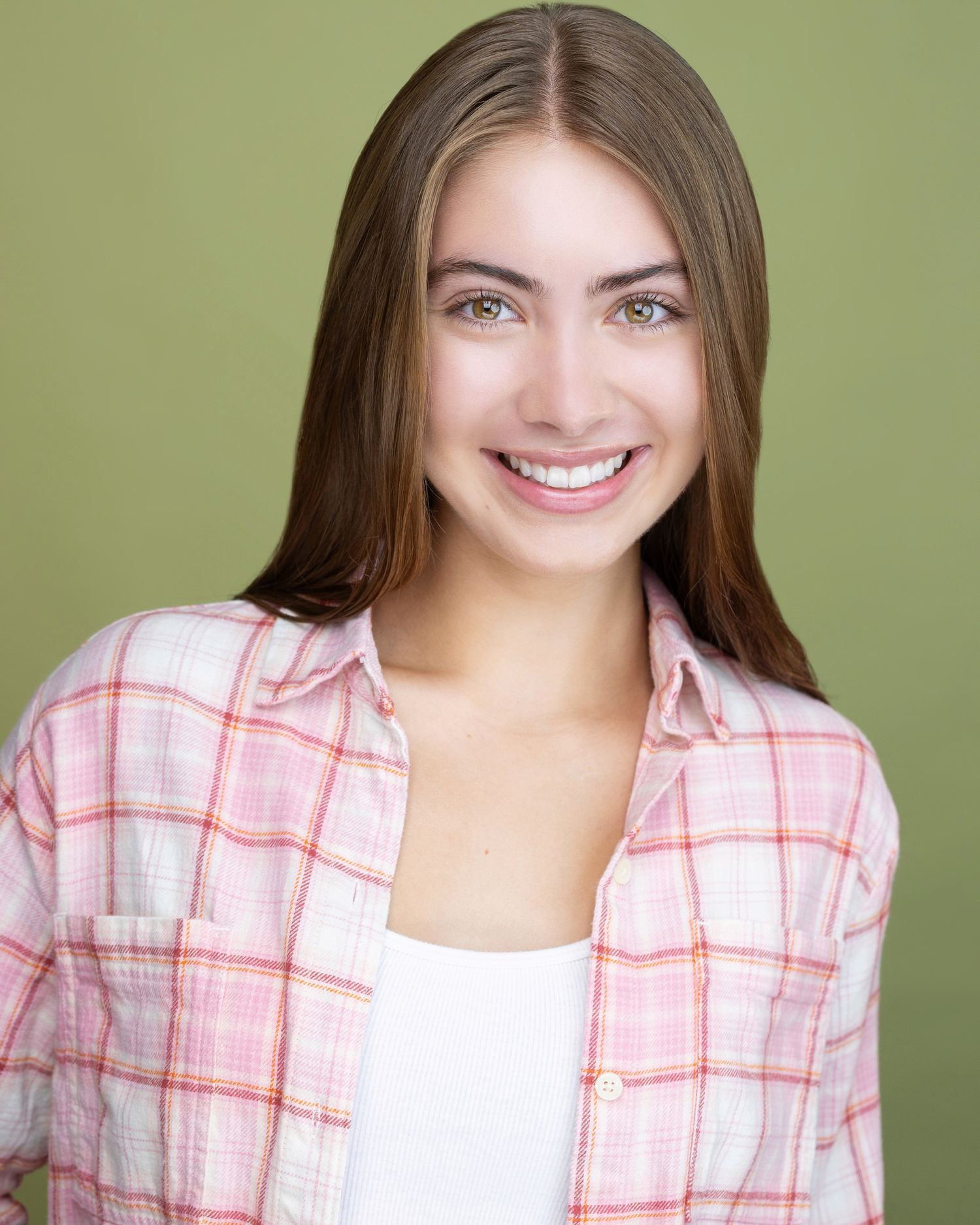 Smiling young woman with long brown hair wearing a pink plaid shirt, posing against a green background.