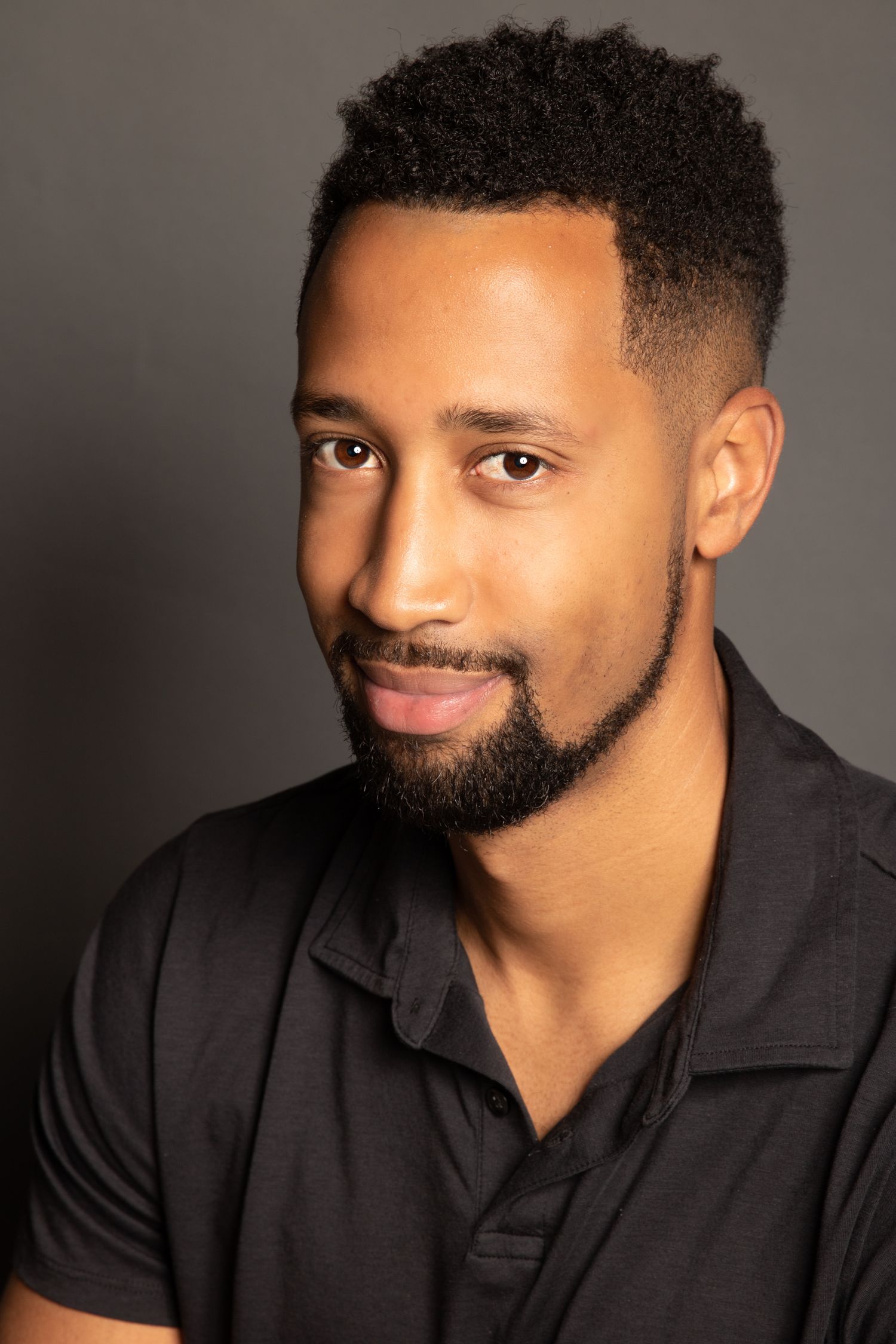 Studio portrait of a person in a black polo shirt with a warm smile and neat beard.