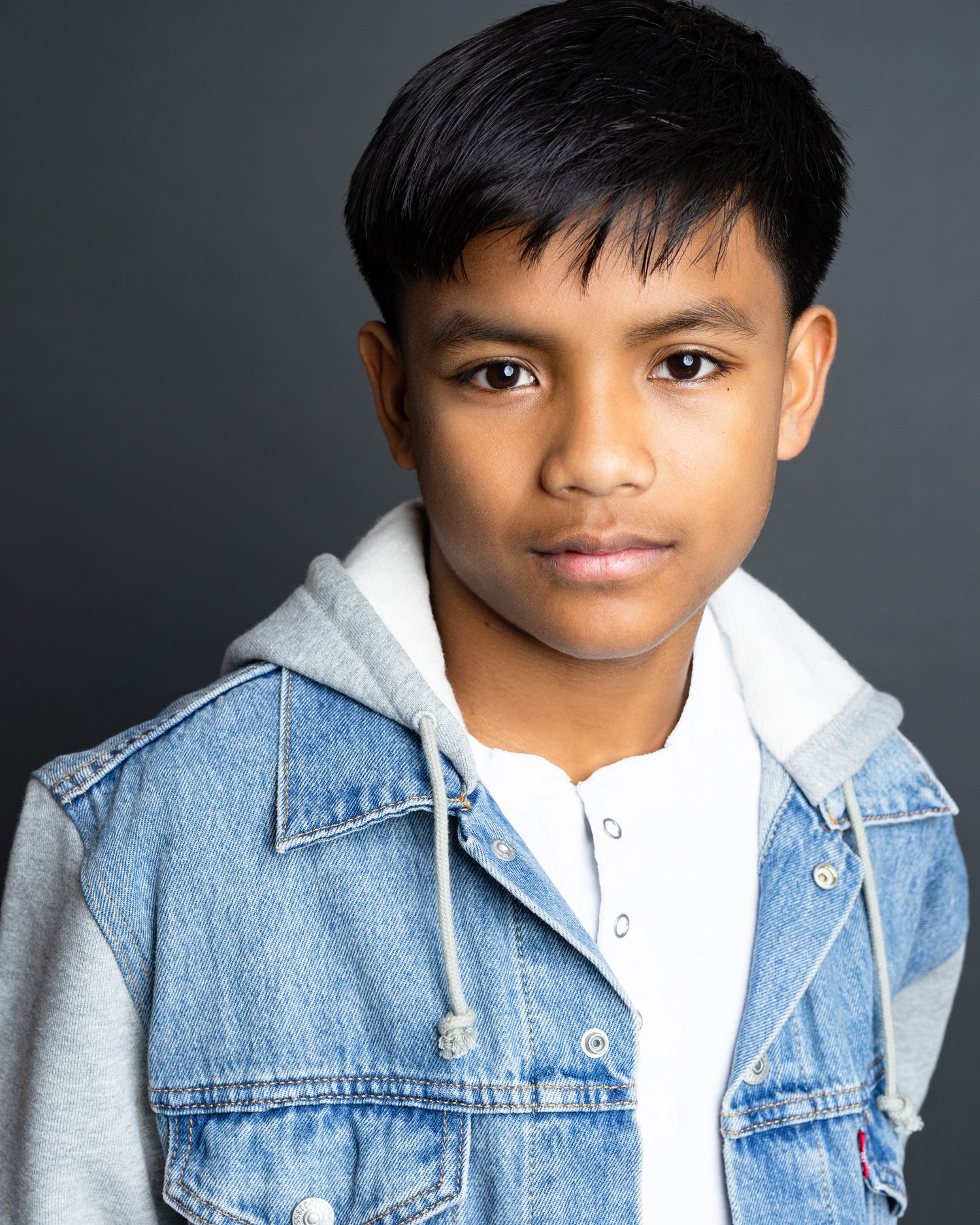 Portrait of a young person wearing a denim jacket over a white hoodie against a dark gray background.