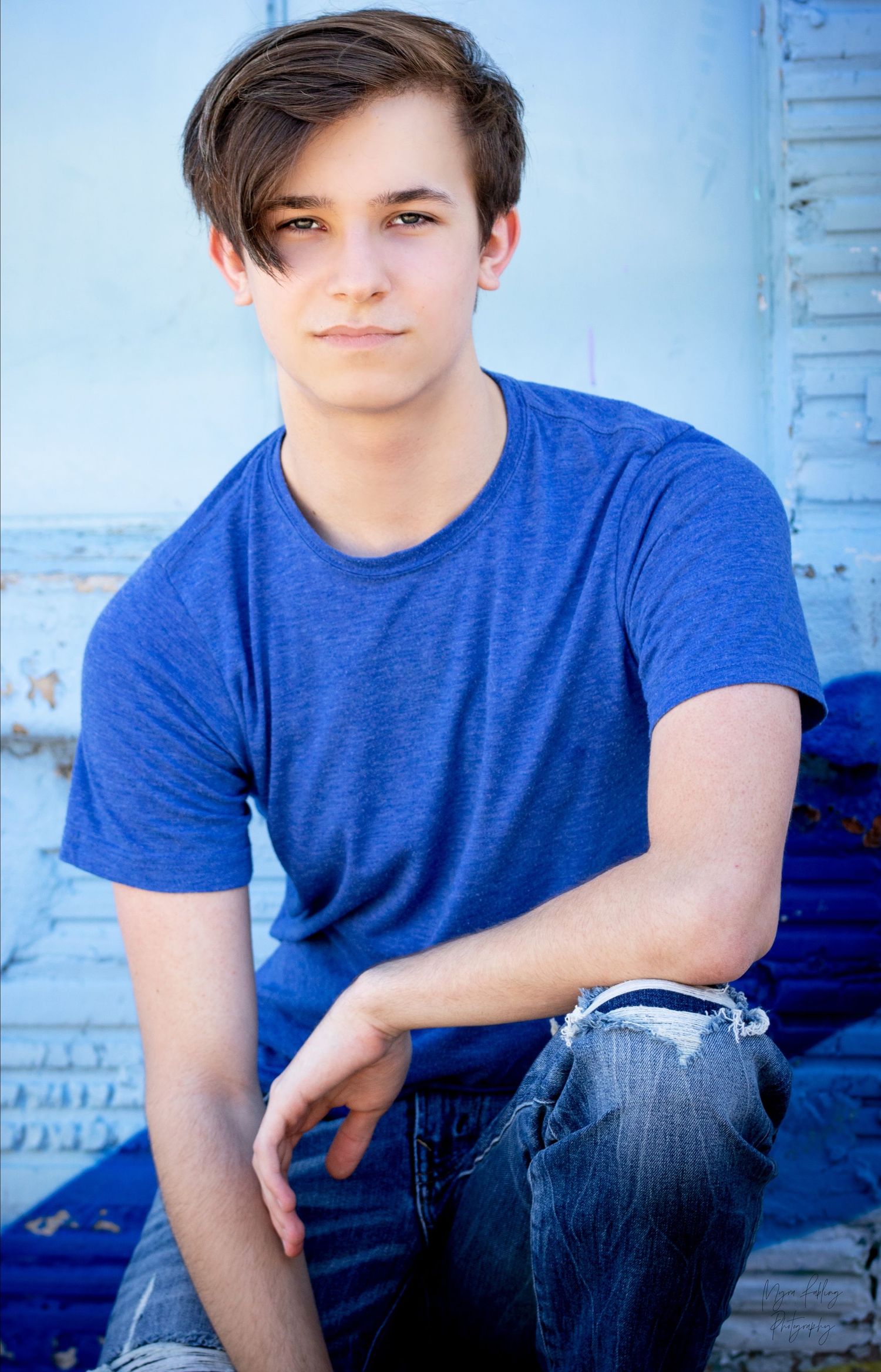Casual outdoor portrait of a young person in blue t-shirt sitting against weathered wall.