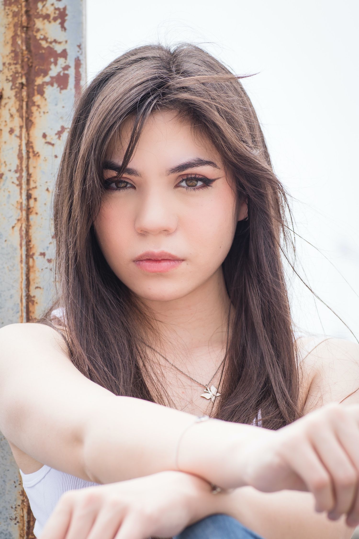 Portrait of a young woman with long dark hair leaning on rustic metal surface.