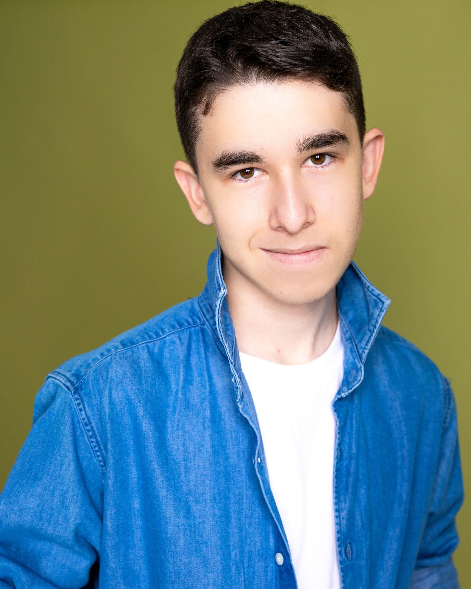 Studio portrait of a young person wearing a denim jacket against an olive green background.