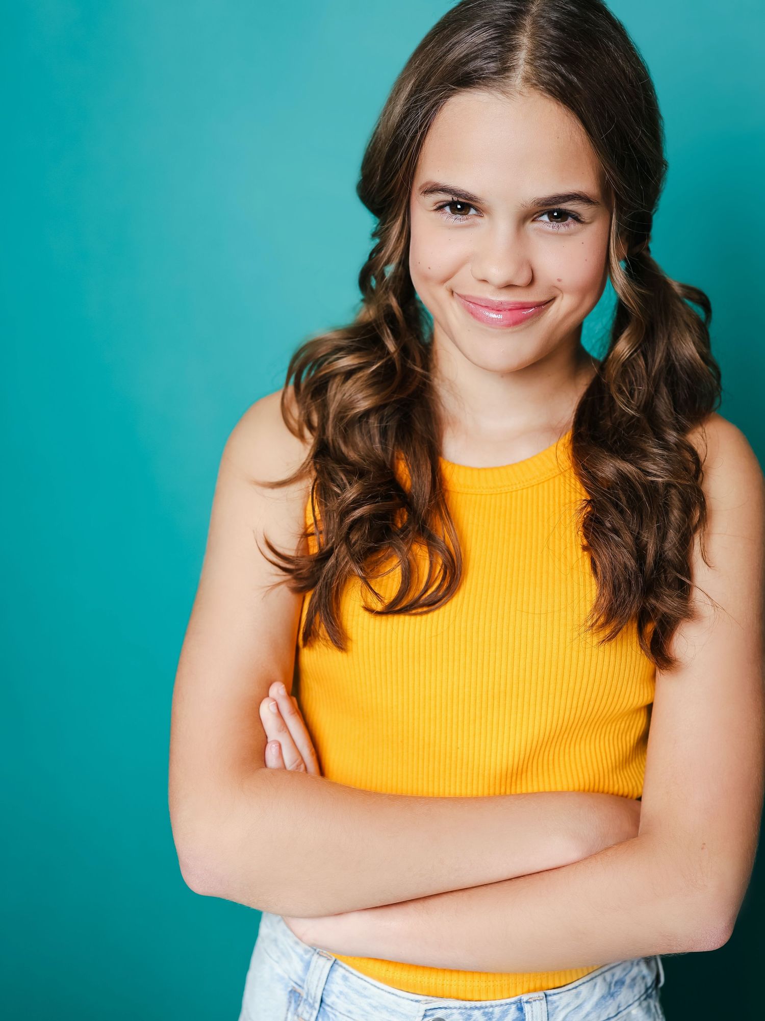 Casual portrait in yellow tank top and denim against turquoise background with crossed arms.