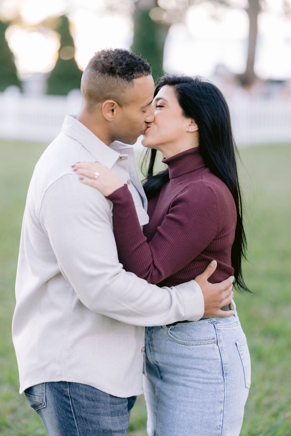 A romantic couple sharing a tender kiss outdoors while wearing casual attire in a park setting.