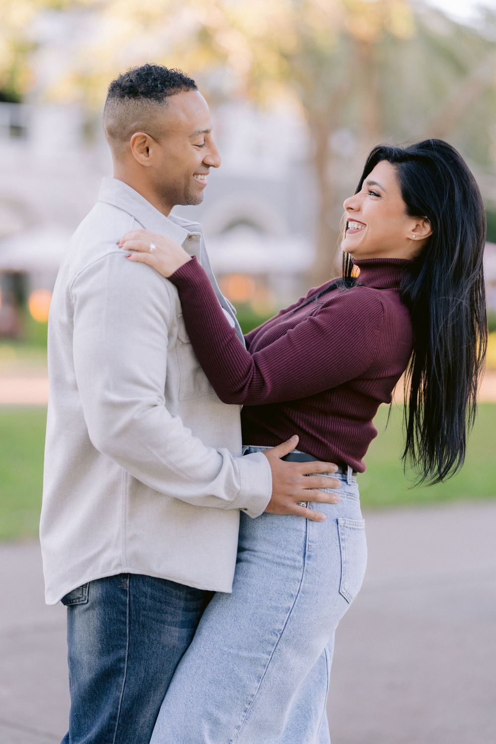 A romantic couple embraces and smiles at each other outdoors while wearing casual autumn clothing.