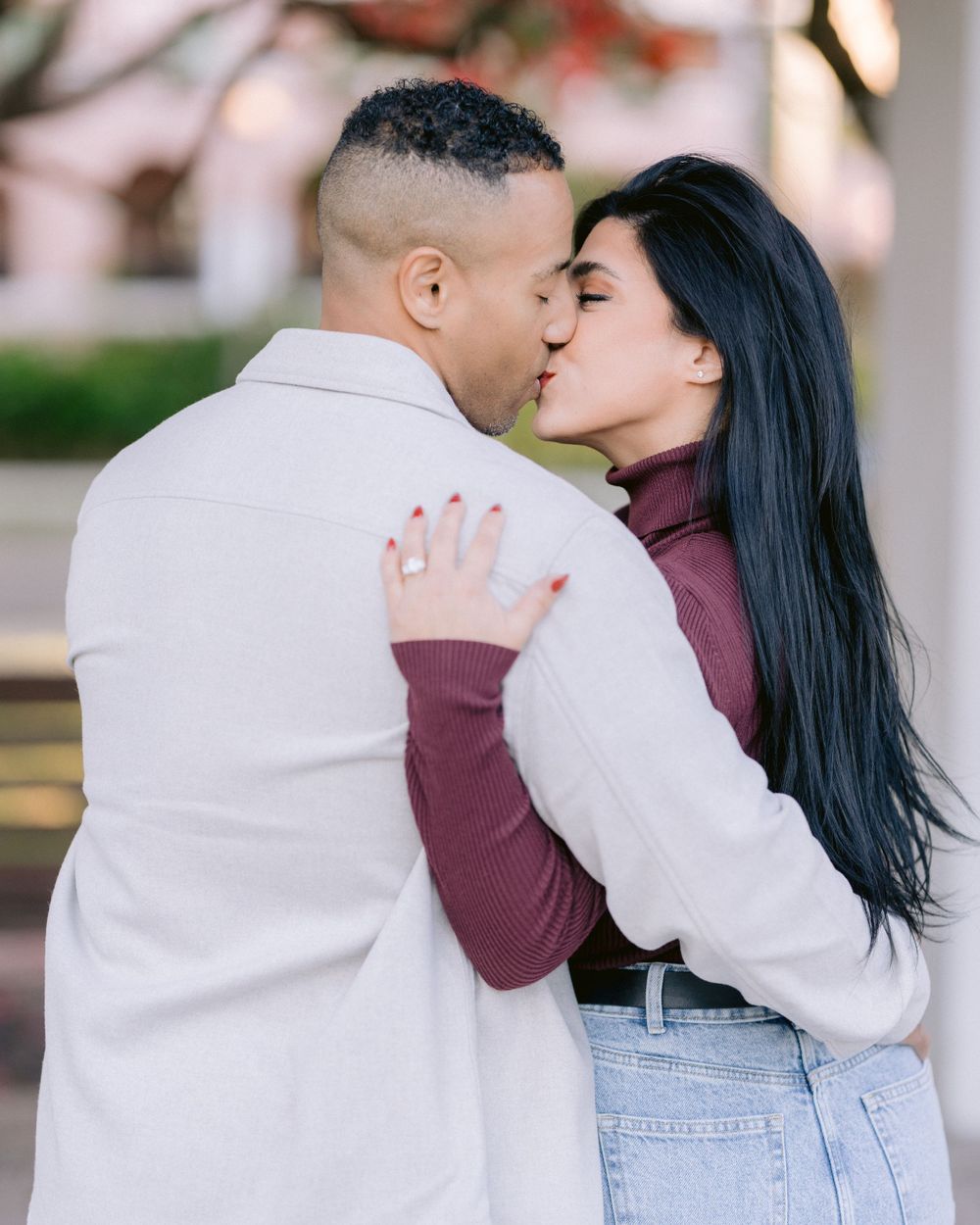 A couple shares an intimate embrace outdoors, with one wearing white and the other in burgundy and denim.