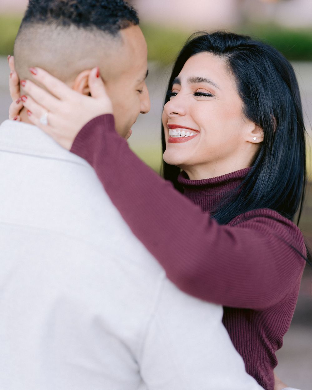 A couple in burgundy and cream sweaters share an intimate moment while smiling outdoors.