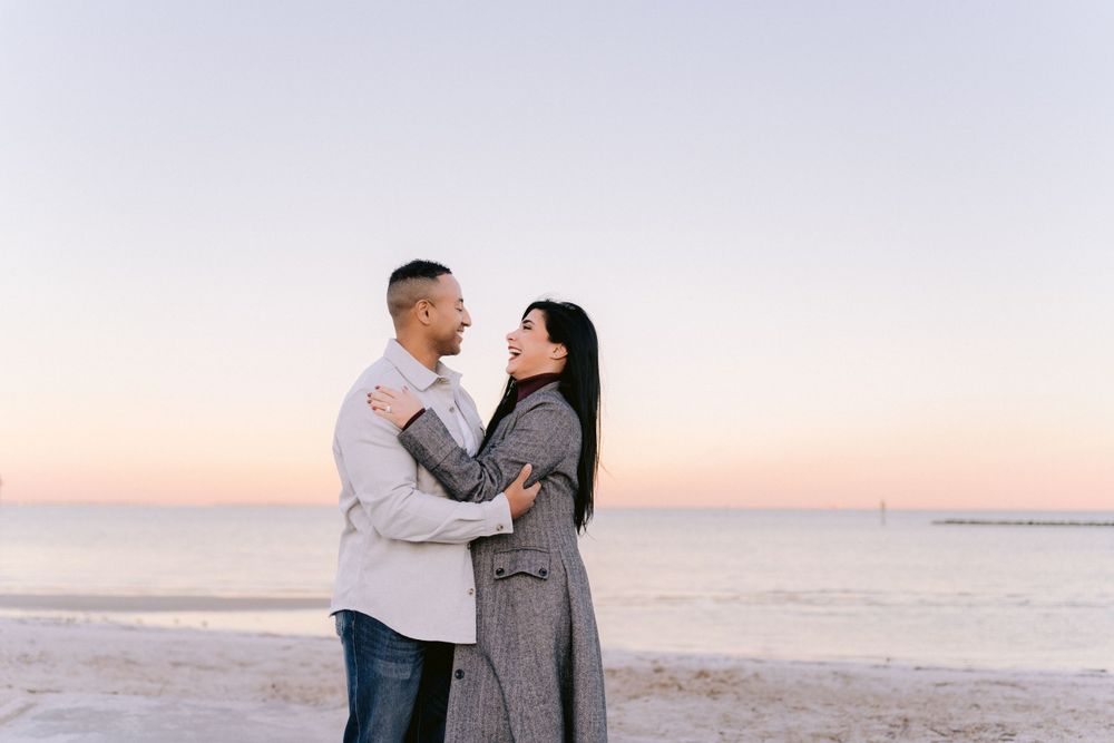 A couple embraces on a beach at sunset with soft pink sky in the background.