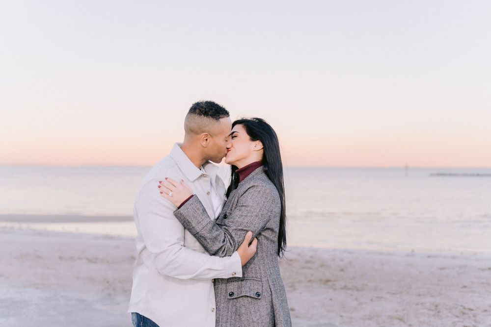 Romantic couple embraces on beach at sunset with pastel sky and calm ocean in background.