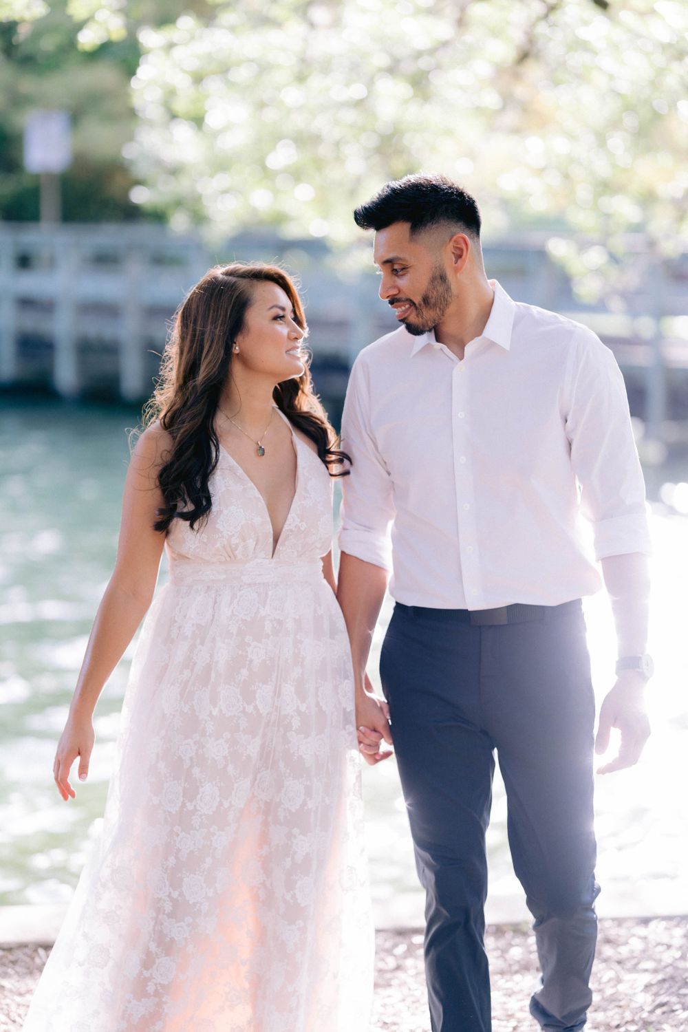 A romantic couple walks together by a waterfront, the woman wearing a flowing white lace dress and man in formal attire.