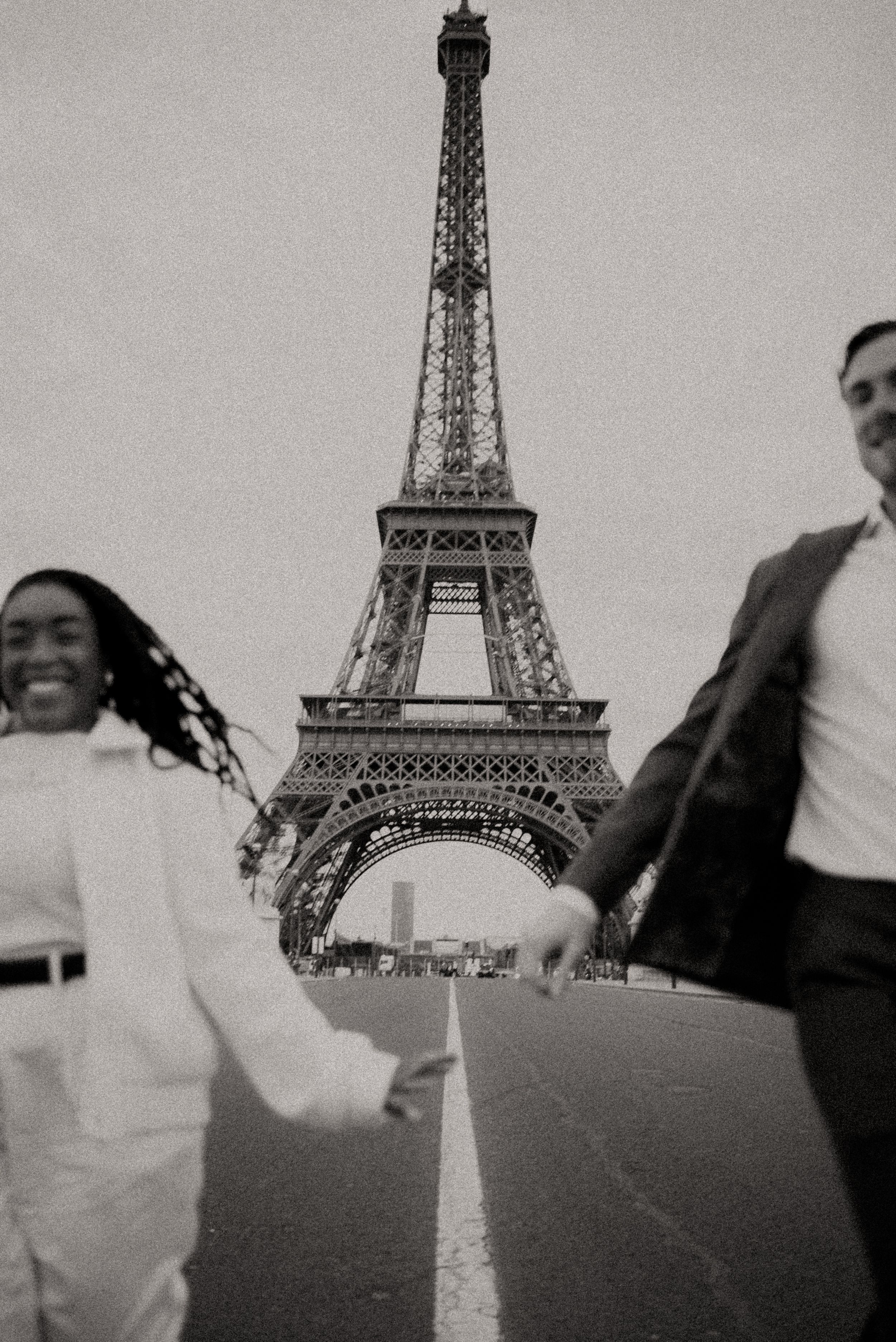 A black and white photo of a couple walking playfully near the Eiffel Tower.