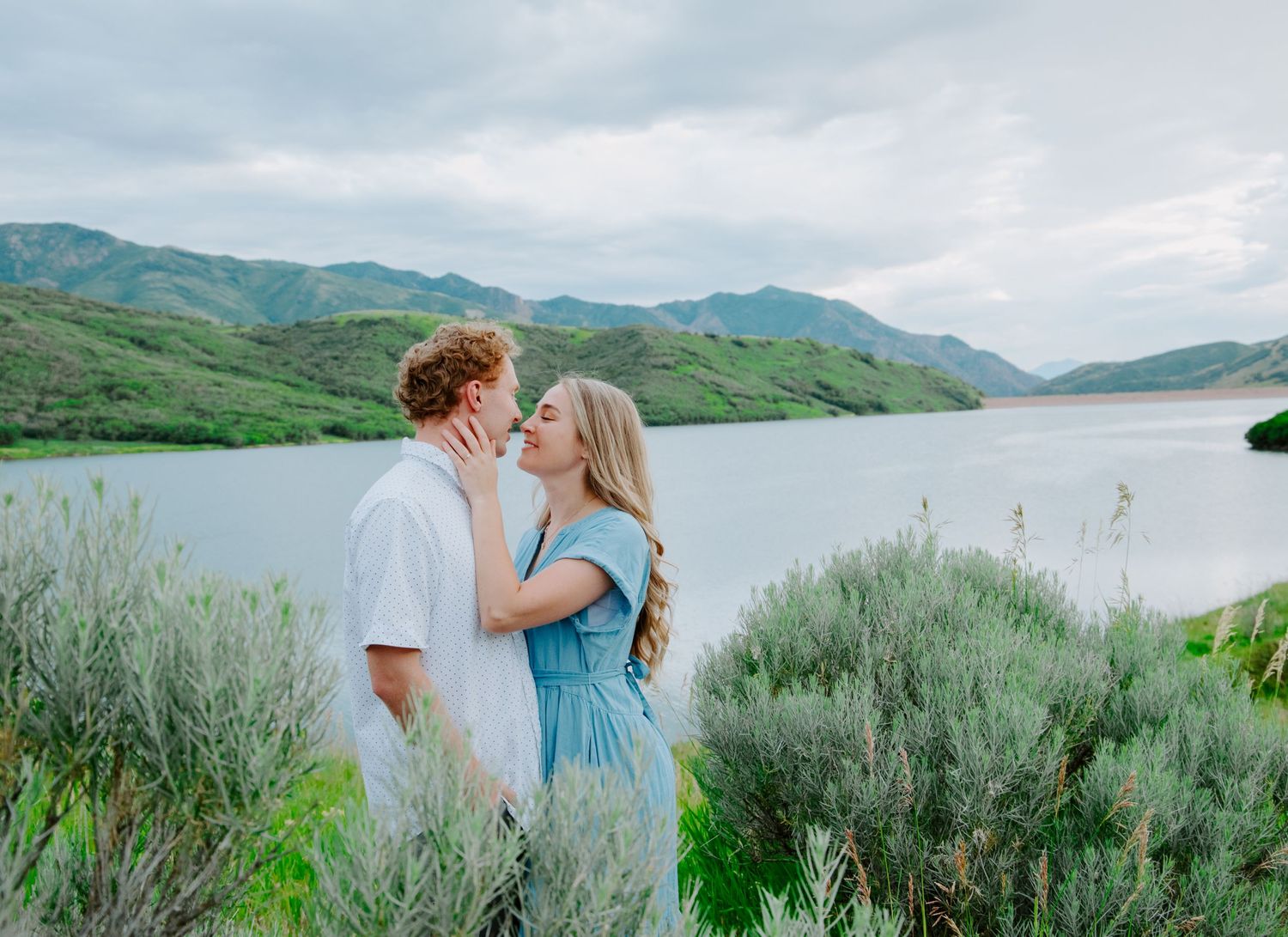 Romantic couple poses together against scenic mountain lake backdrop with green foliage.