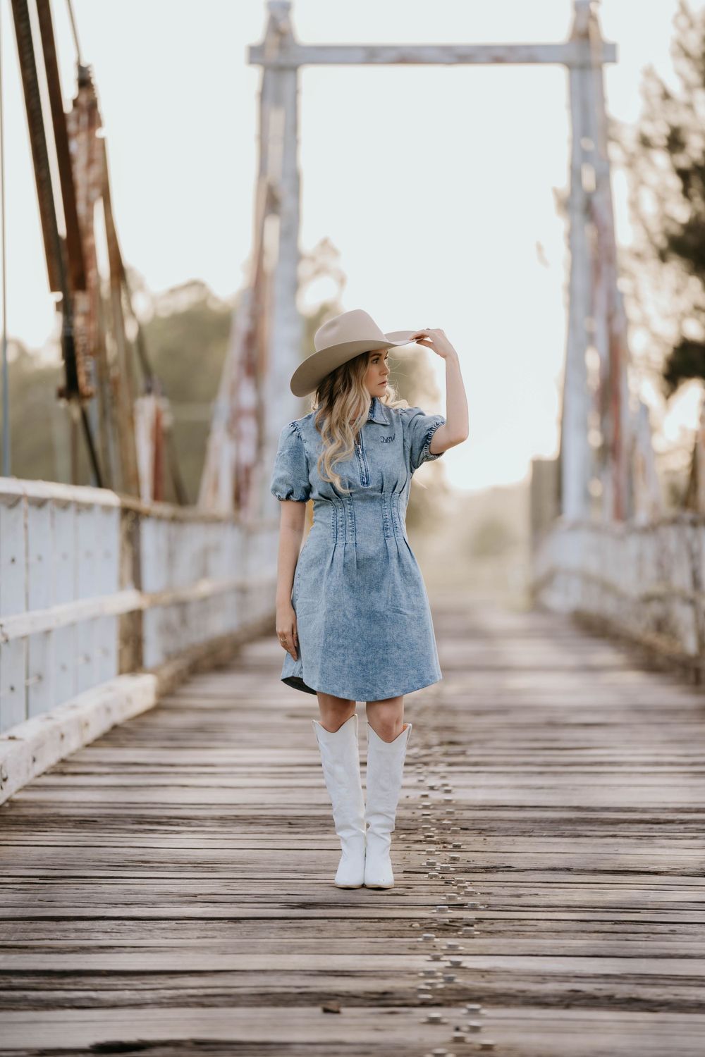 Person in denim dress and white cowboy boots stands on wooden bridge during golden hour sunset.