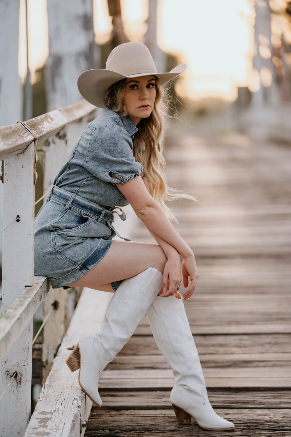 A person in a denim dress and tan hat poses on a wooden boardwalk wearing white cowboy boots at sunset.