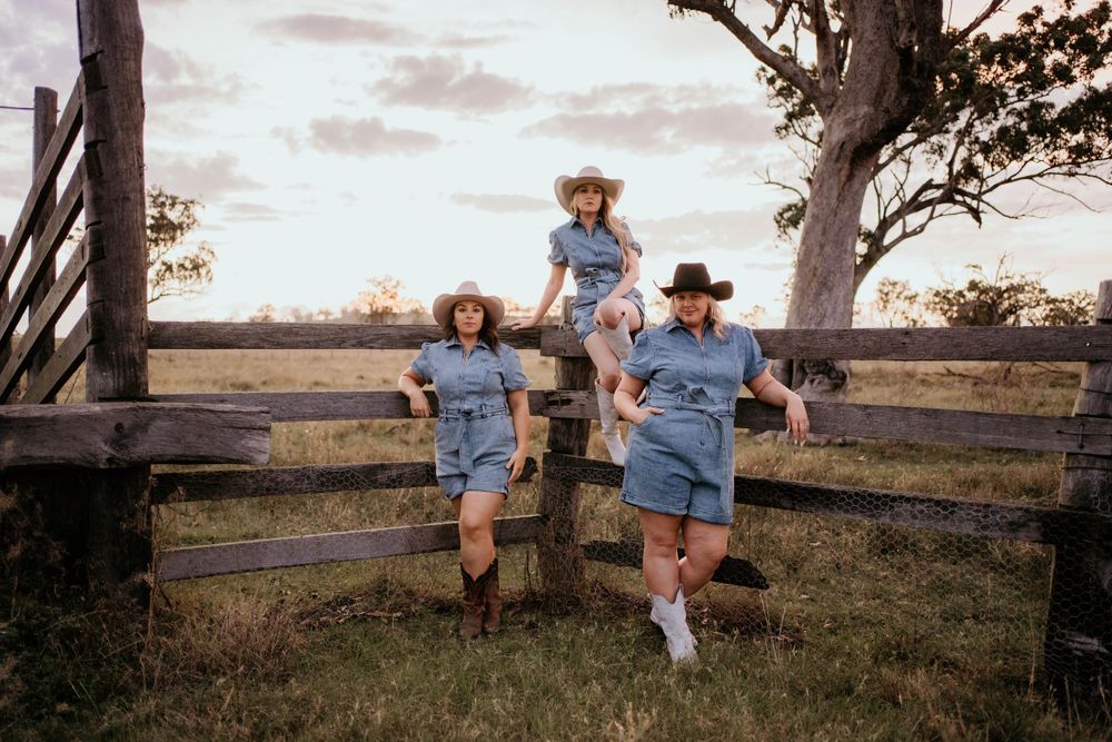 Three people in matching blue outfits pose on a rustic wooden fence during sunset in a rural setting.