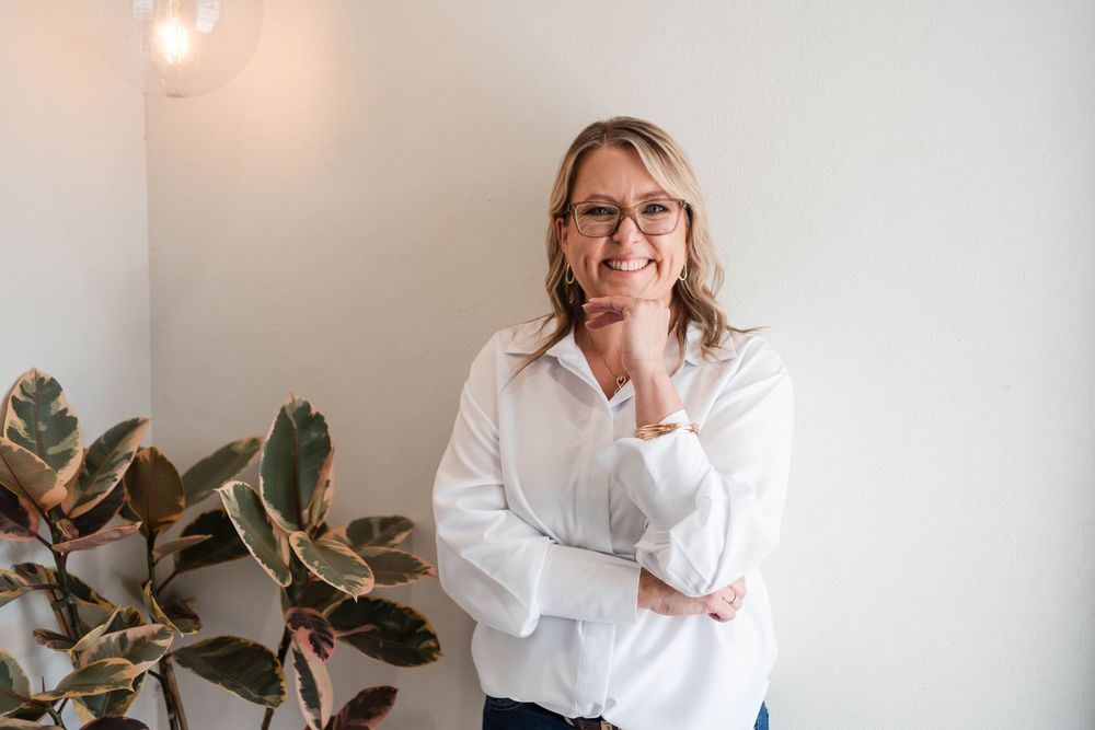 A person in a white button-down shirt smiles while standing next to a potted plant against a light gray wall.