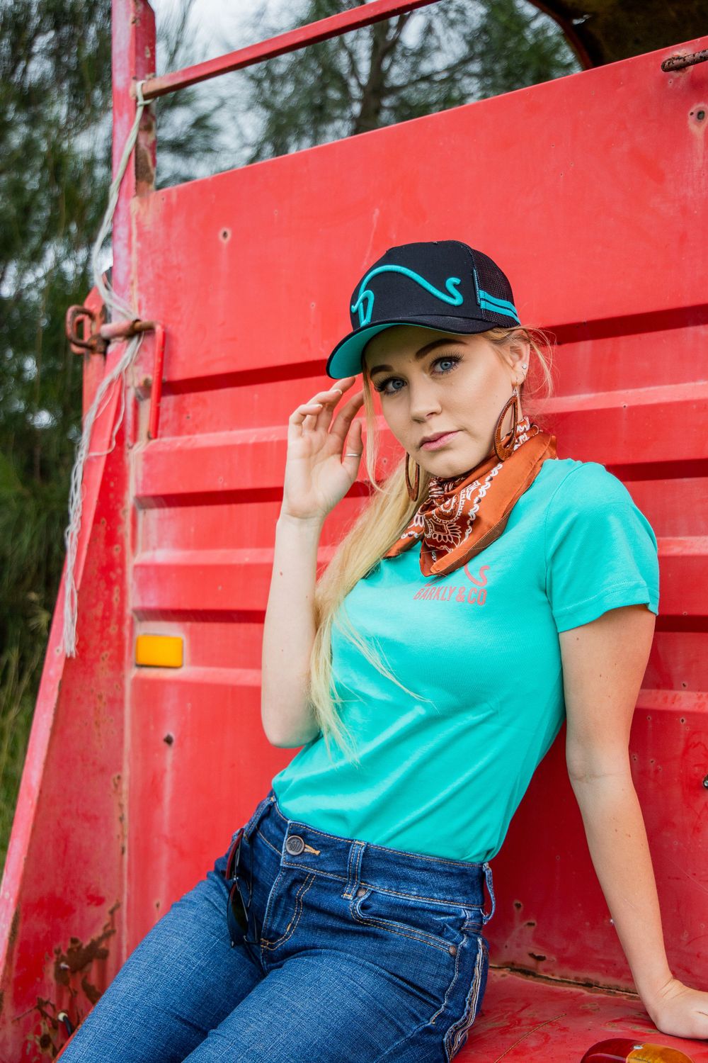 A western style outfit with turquoise shirt and baseball cap posed against a red metal structure.