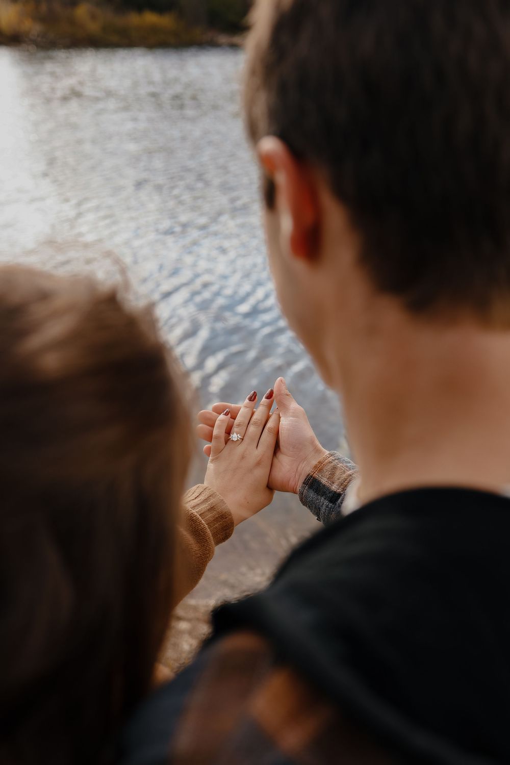 A couple holds hands while looking out at a peaceful lake, showcasing their wedding rings.
