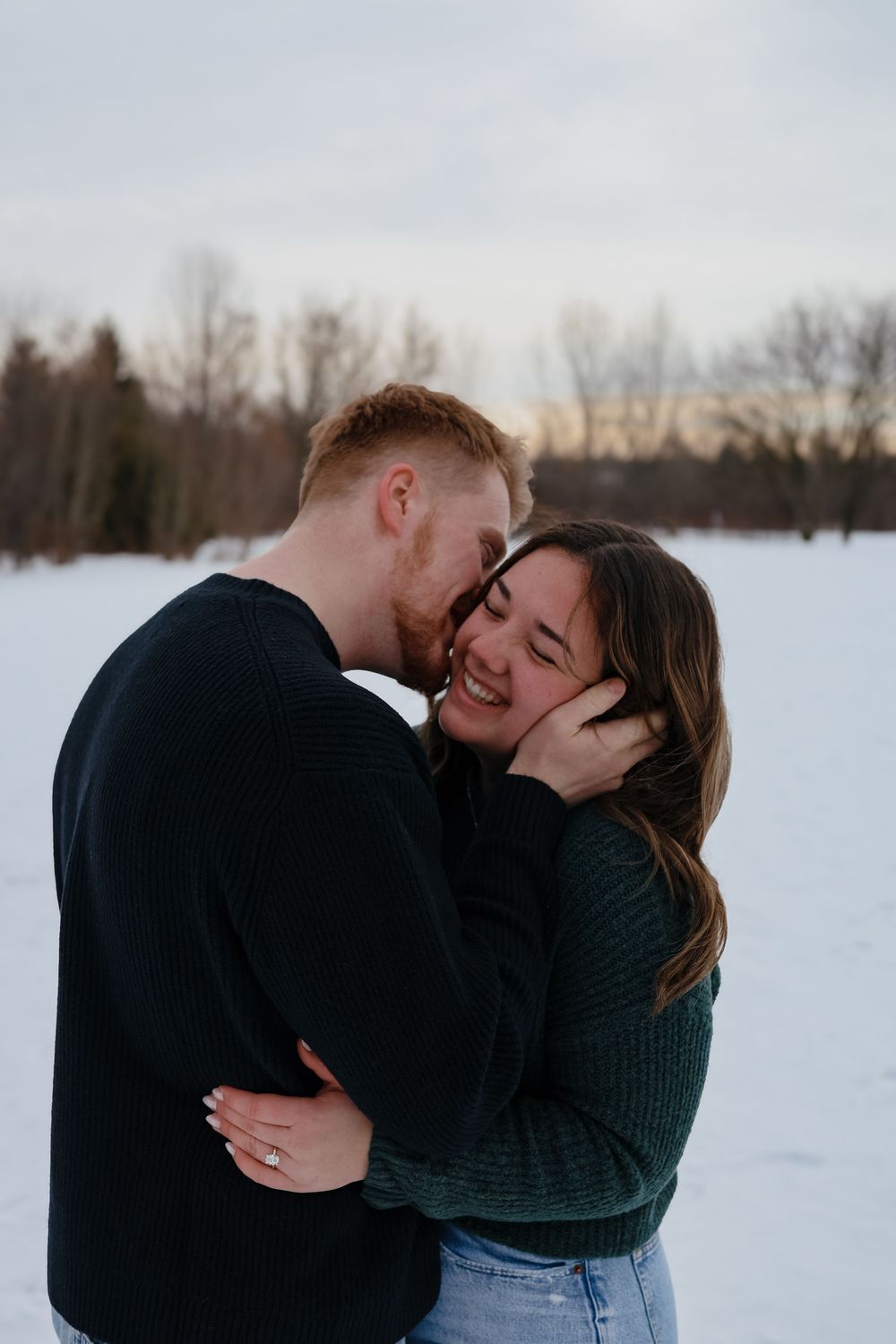 A couple shares an intimate moment while embracing in a snowy winter landscape at dusk.