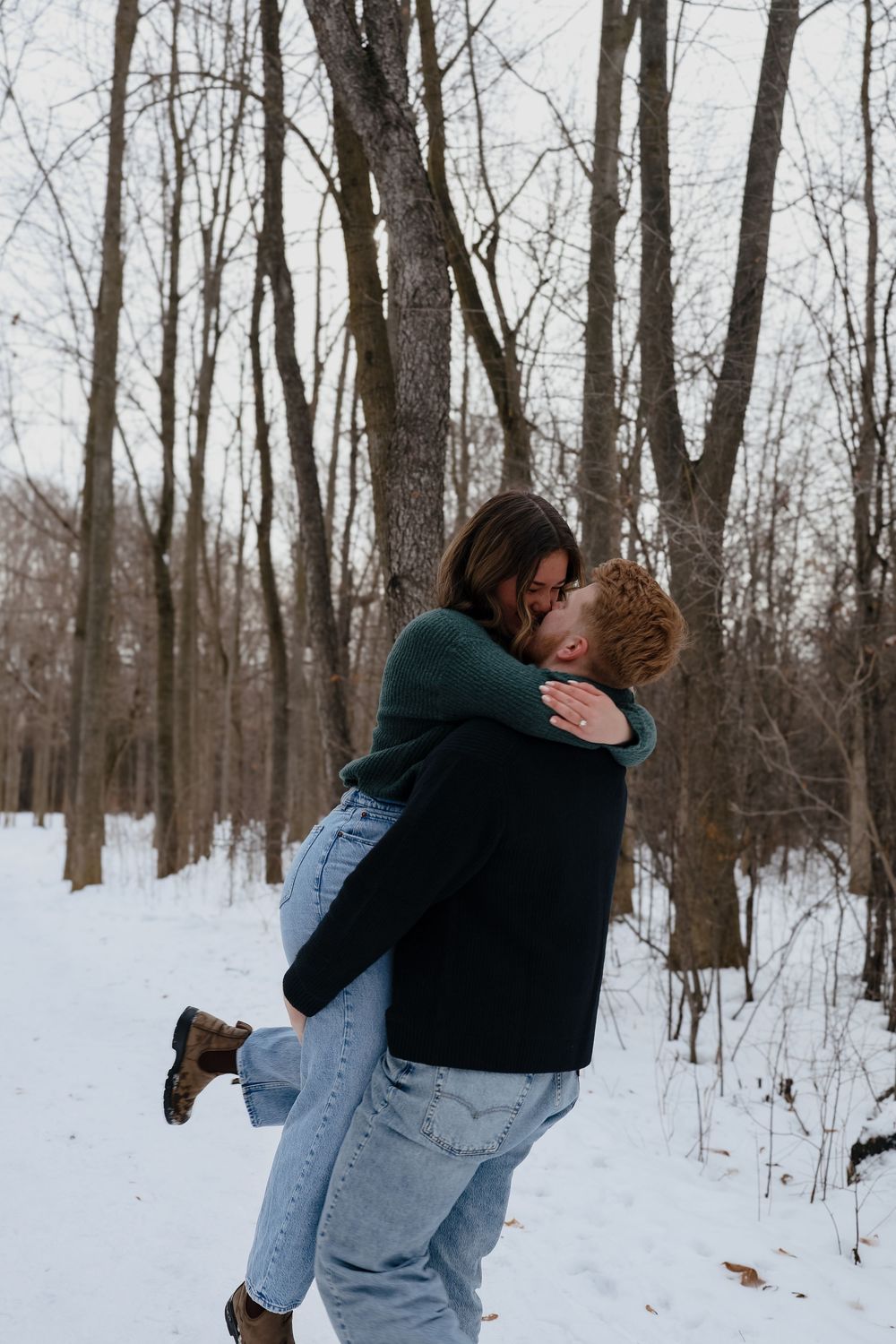 A couple embraces in a snowy winter forest setting with bare trees in the background.