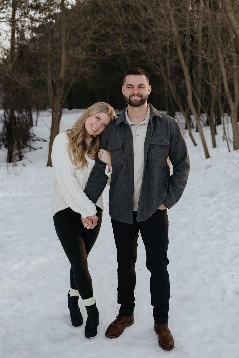 A couple poses together in a snowy winter forest setting wearing coordinated casual outfits.