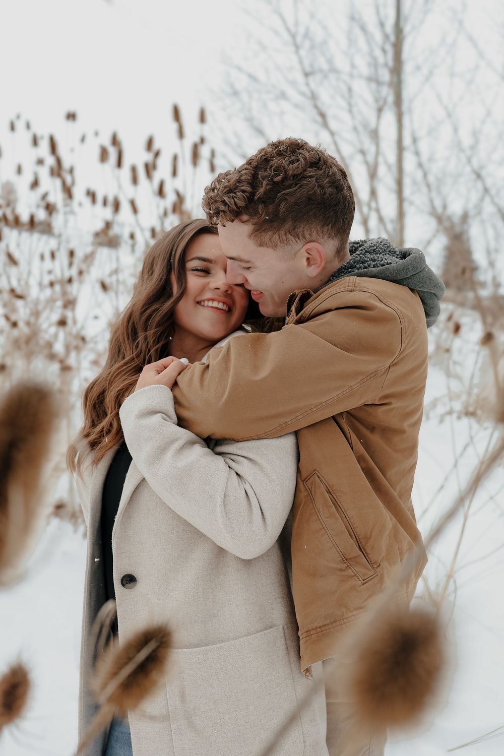 A romantic couple shares an intimate winter moment outdoors wearing beige and brown coats in the snow.